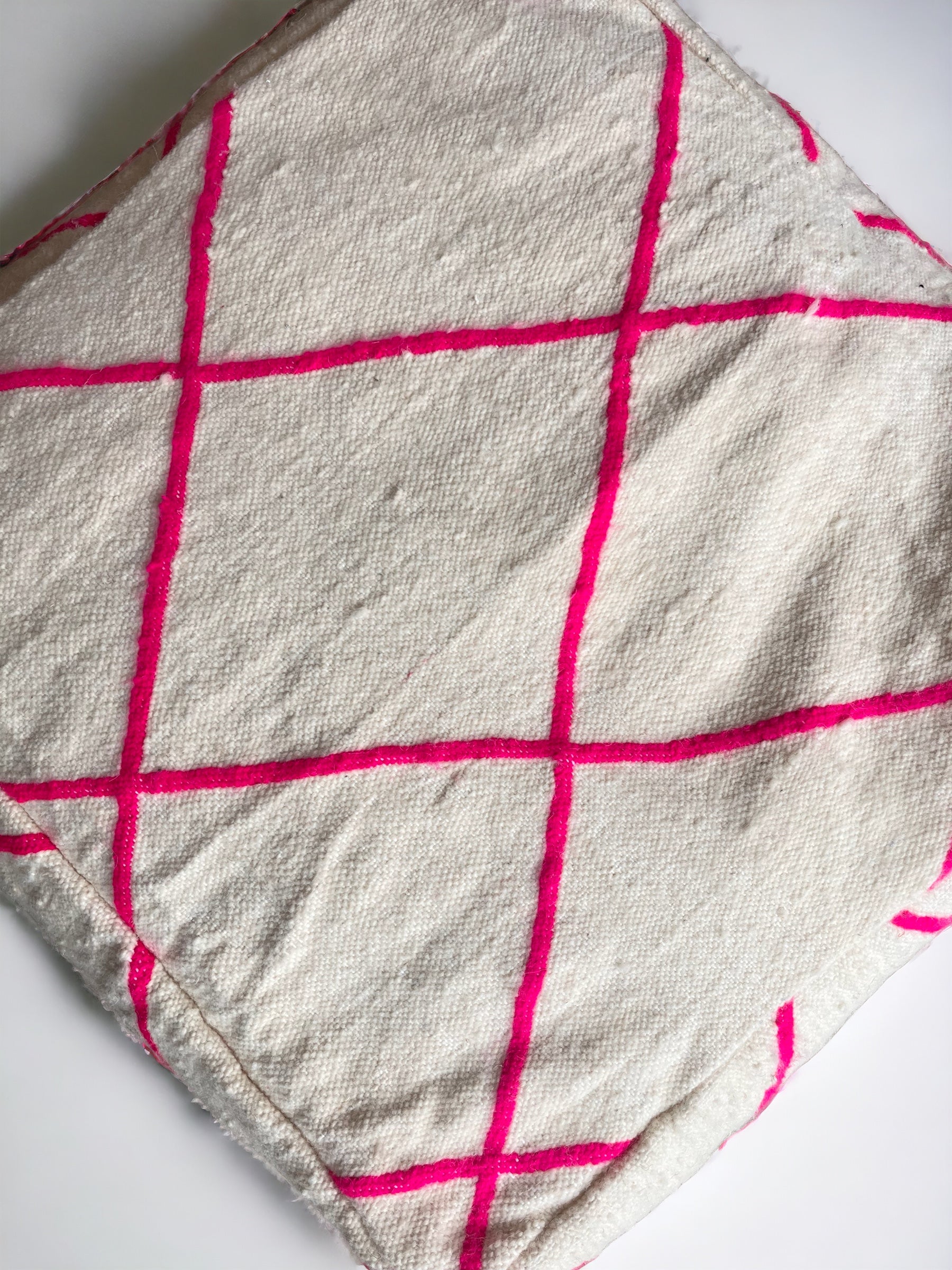 Das Bodenkissen - Mina von LEY! zeigt cremefarbene, strukturierte Schurwolle mit sich kreuzenden, pinkfarbenen Linien, die große Quadrate bilden. Es ist ideal für Handarbeitsprojekte und wird gefaltet auf weißem Hintergrund gezeigt.