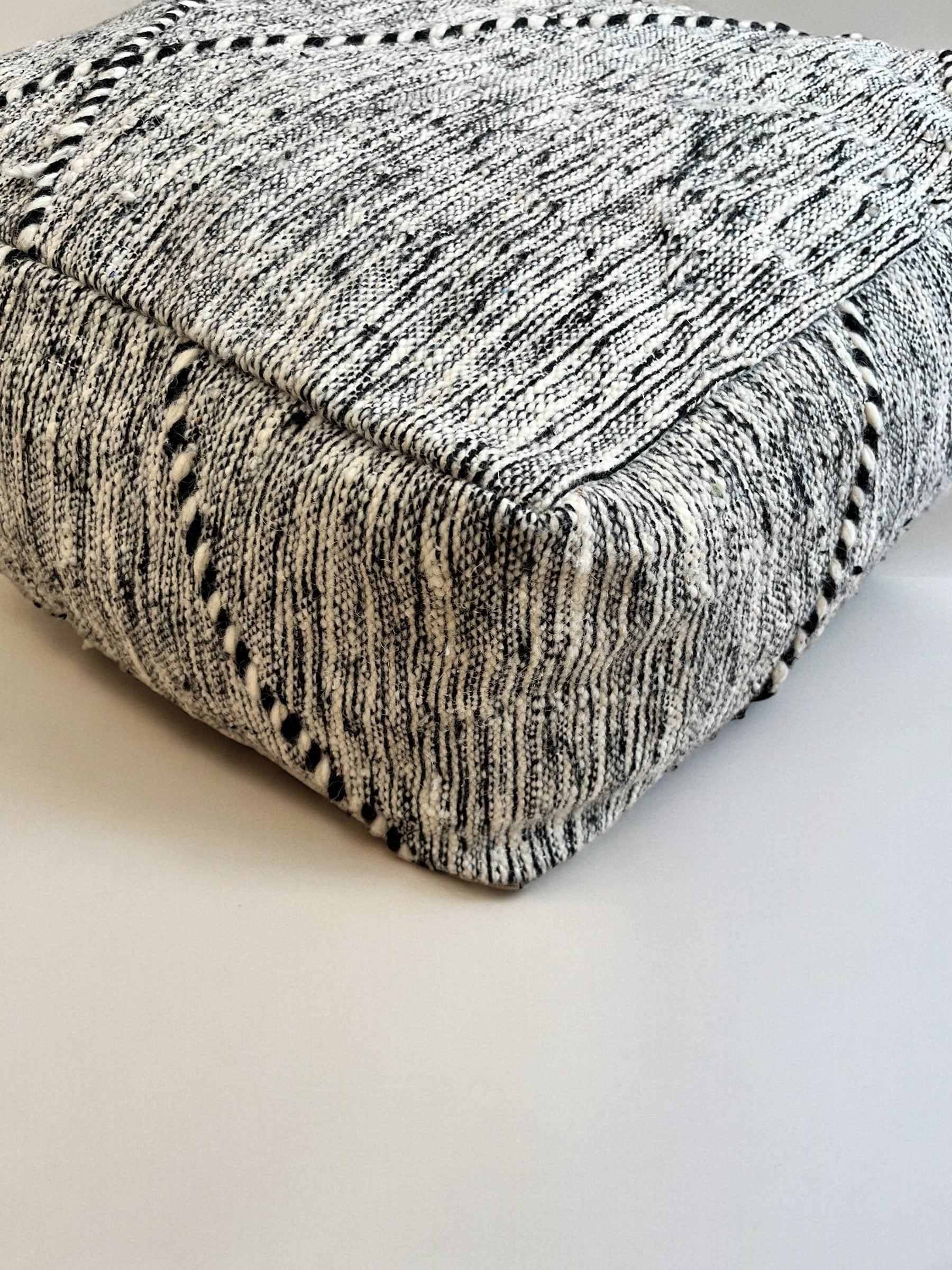 Eine Nahaufnahme des quadratischen Kleinen Bodenkissens - Mica Mini von LEY!, handgewebt in einem schwarz-weißen Muster mit schwarzen Steppnähten, ausgestellt auf einer hellen Fläche.