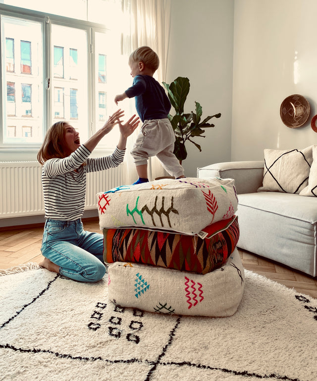 Bodenkissen auf dem Teppich in einem Wohnzimmer vor einem Sofa. Eine Frau fängt ihr Kind das von Bodenkissen hüpft.