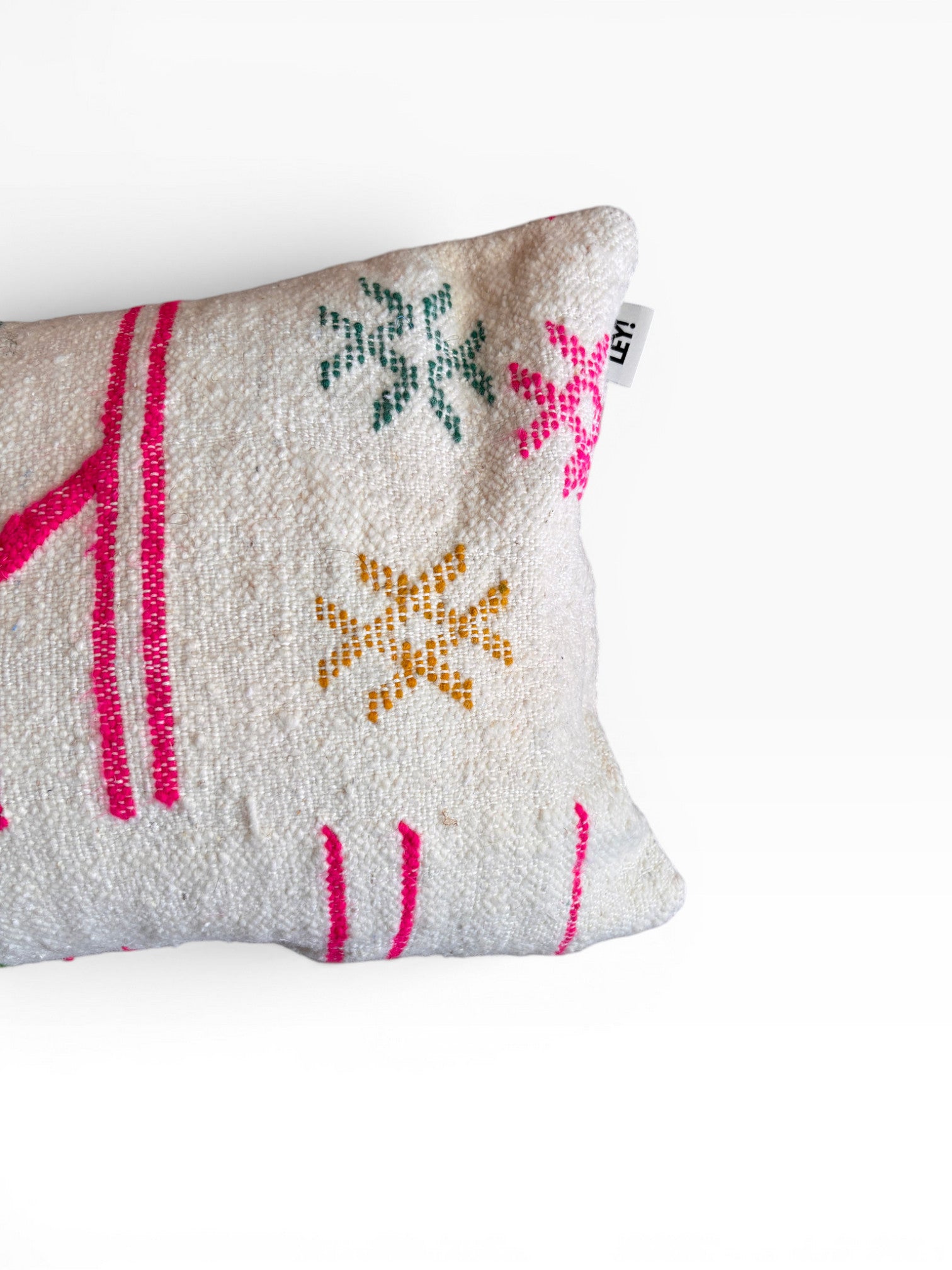 Das LEY! Kissen - Colorful Line - 40 x 60 von LEY! ist ein rechteckiges weißes Kissen mit rosa, grünen und gelben gestickten Stern- und Linienmotiven. Es ist mit einem kleinen Stoffanhänger am Rand versehen, der teilweise auf einem unifarbenen Hintergrund abgebildet ist.