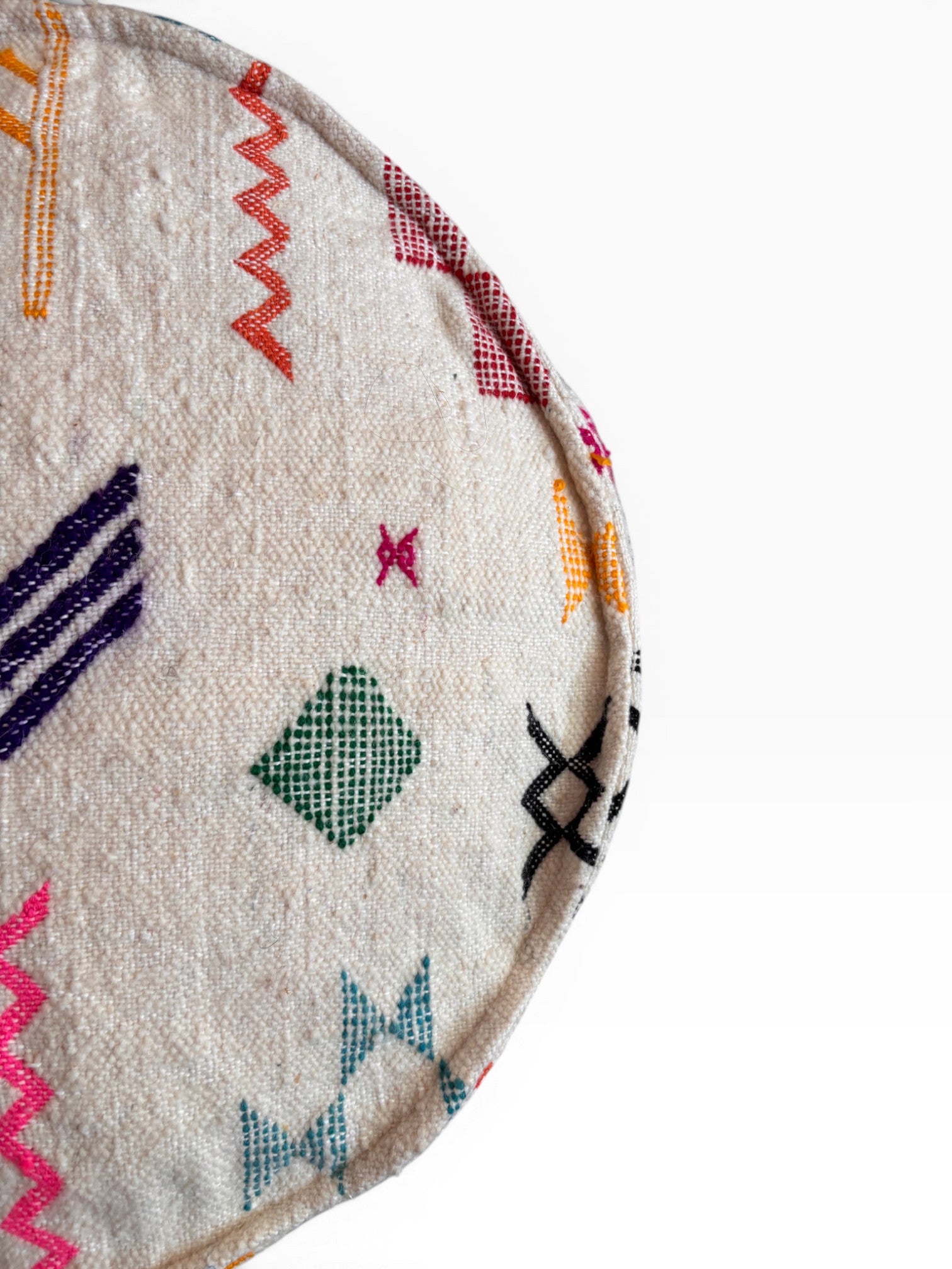 Eine Nahaufnahme des LEY! Bodenkissen - Colorful Line Round - Tafika zeigt strukturierte Schurwolle mit grüner, orangefarbener, rosa, schwarzer und lila geometrischer Stickerei auf cremefarbenem Untergrund; ein Teil dieses Boho-Kissens erscheint vor einem schlichten weißen Hintergrund.