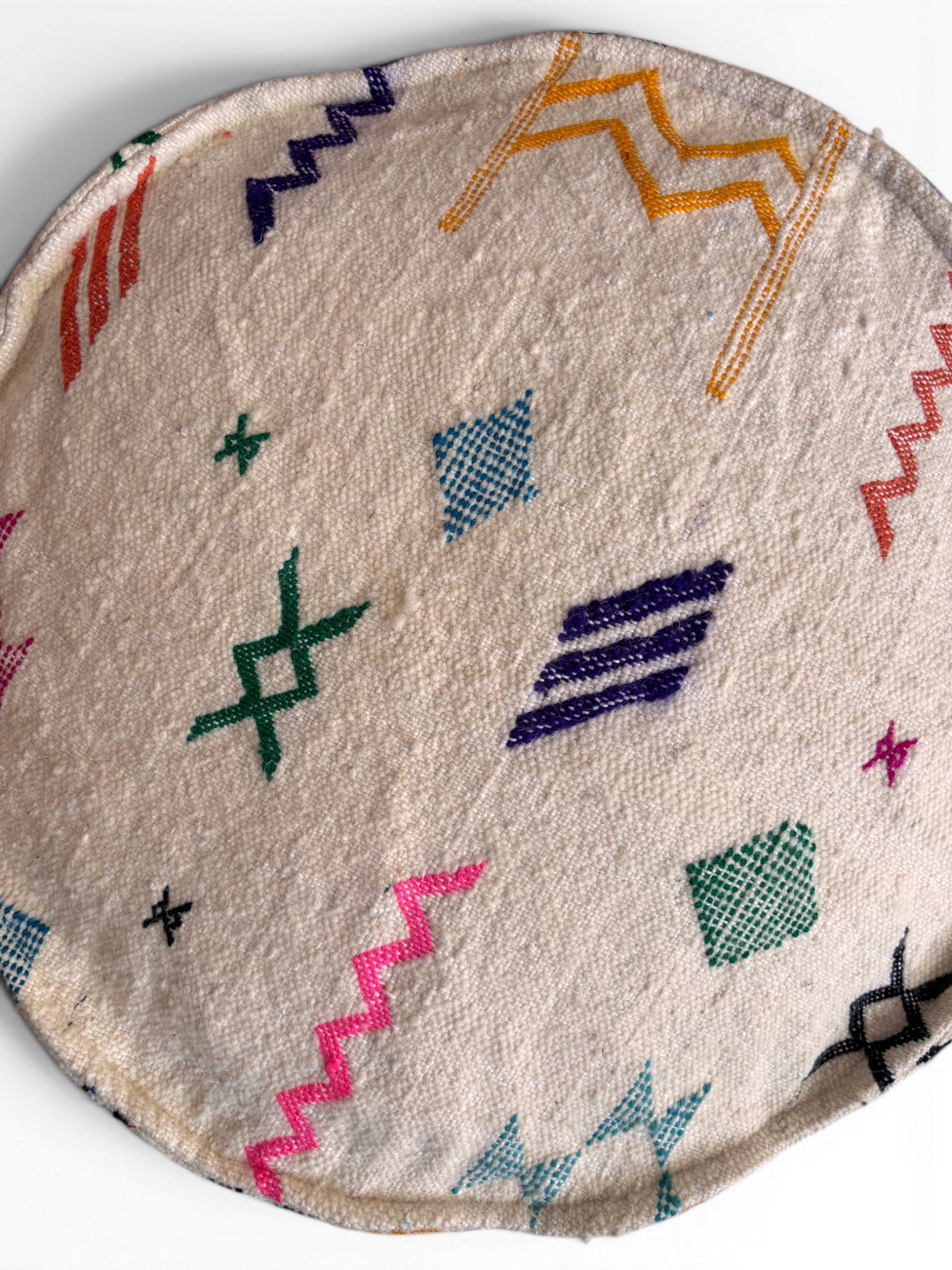 Das LEY! Bodenkissen - Colorful Line Round - Tafika ist ein rundes, beigefarbenes Sitzkissen aus Schurwolle mit lebhaften geometrischen Stickereien in Grün, Lila, Blau, Gelb, Rosa, Rot und Schwarz auf weißem Untergrund.