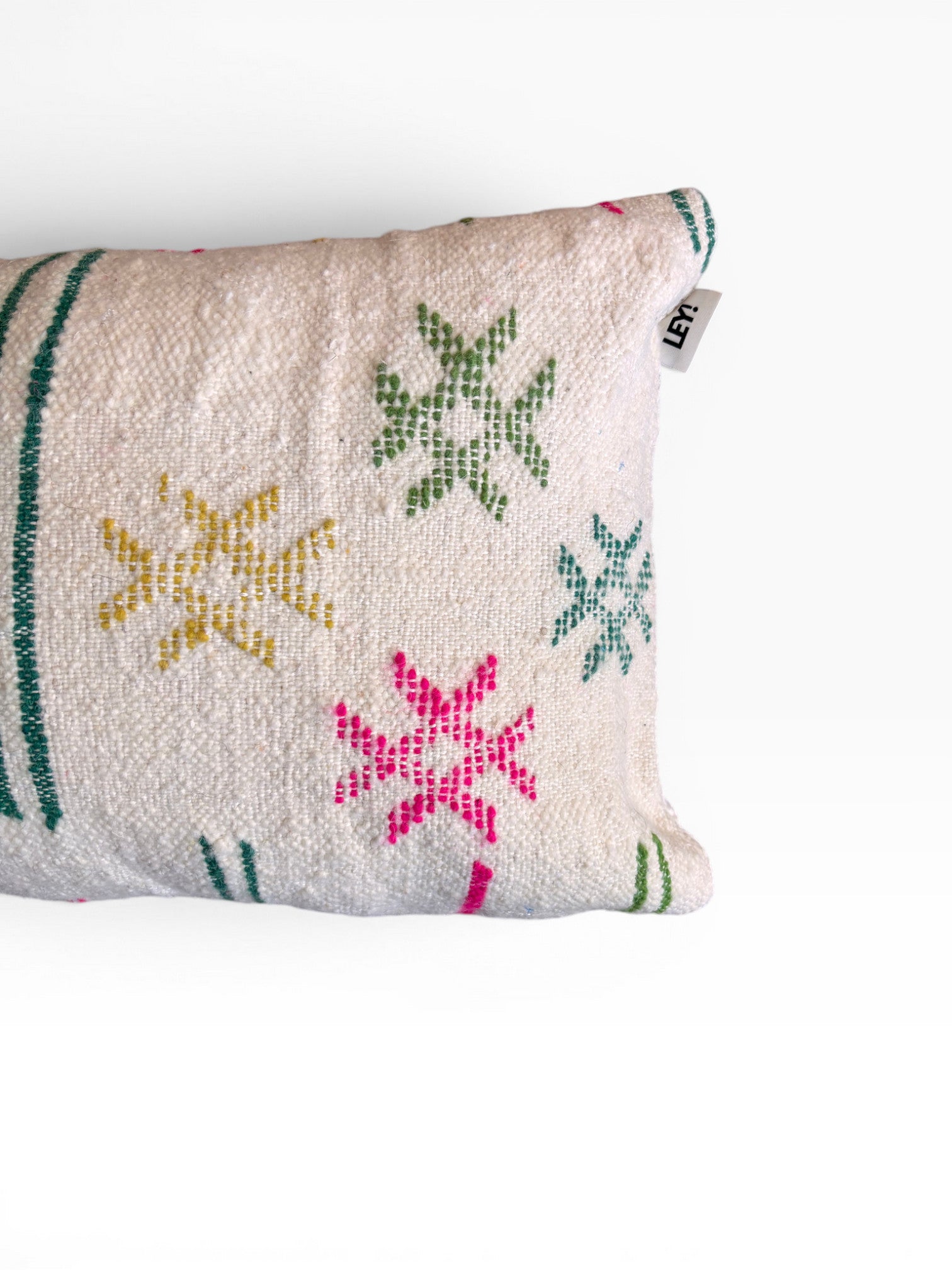 Das LEY! Kissen - Colorful Line (40 x 60) von LEY! ist ein weißes, strukturiertes Kissen mit rosa, gelben und grünen sternförmigen Mustern und Streifen, die teilweise auf einem einfarbig weißen Hintergrund abgebildet sind. Am Rand ist ein kleines Stoffschildchen angebracht.