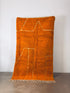 Der LEY! Berber Teppich -Sefa- (1,45 x 2,50) ist ein rechteckiger, orangefarbener, handgefertigter Schurwollteppich mit dicker, zotteliger Textur, beigen, abstrakten Linienmustern und kurzen Fransen am unteren Rand, der vertikal an einer weißen Wand hängt.