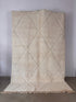 Der BERBER TEPPICH "JUNAH" von LEY! (2,00 x 3,00 m), ein großer cremefarbener Teppich mit einem subtilen Rautenmuster und ausgefransten Rändern, ist aufrecht an einer schlichten Wand auf einem Betonboden abgebildet.