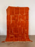 Der LEY! Berber Teppich - Mumina (1,55 x 2,55) ist ein leuchtend orangefarbener, handgefertigter Teppich aus Schurwolle mit leichten geometrischen Mustern, der aufrecht vor einer weißen Wand gezeigt wird. Er hat unten Fransen und eine leicht unebene Oberkante.