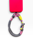 Ein Handy mit einer rosa Hülle, einer LEY! Schwarz-Weiß „LUJA“-Handykette sowie einem farbenfrohen Handyband.