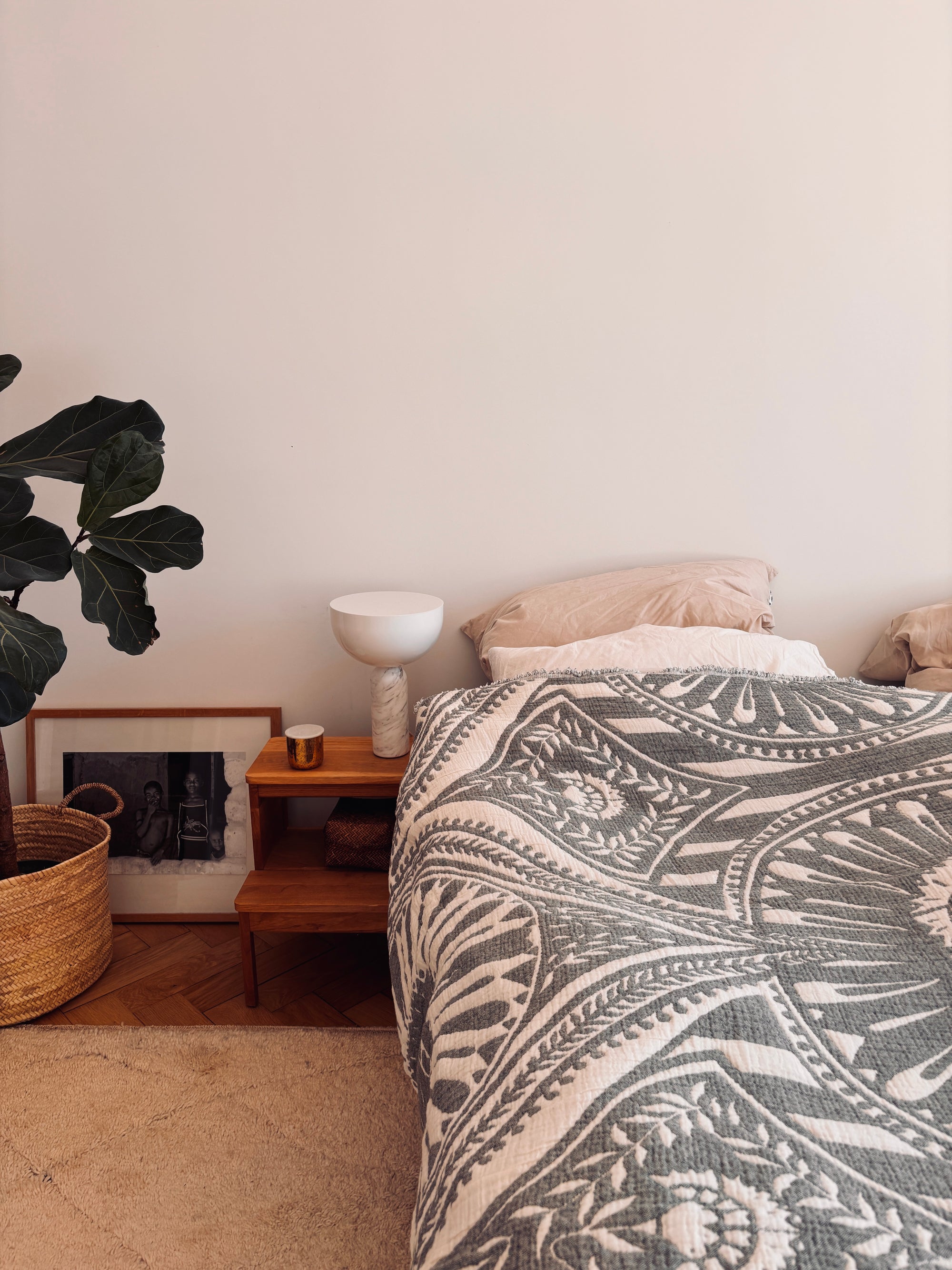 In einer gemütlichen Schlafecke steht eine LEY! CALMA Musselin Decke in Dunkelgrau (2,20 x 2,30 m) auf dem Bett, mit einem hölzernen Nachttisch, einer Lampe, einer Tasse, einer Topfpflanze, einem Weidenkorb, einem Bilderrahmen und einem beigen Teppich auf dem Holzboden.