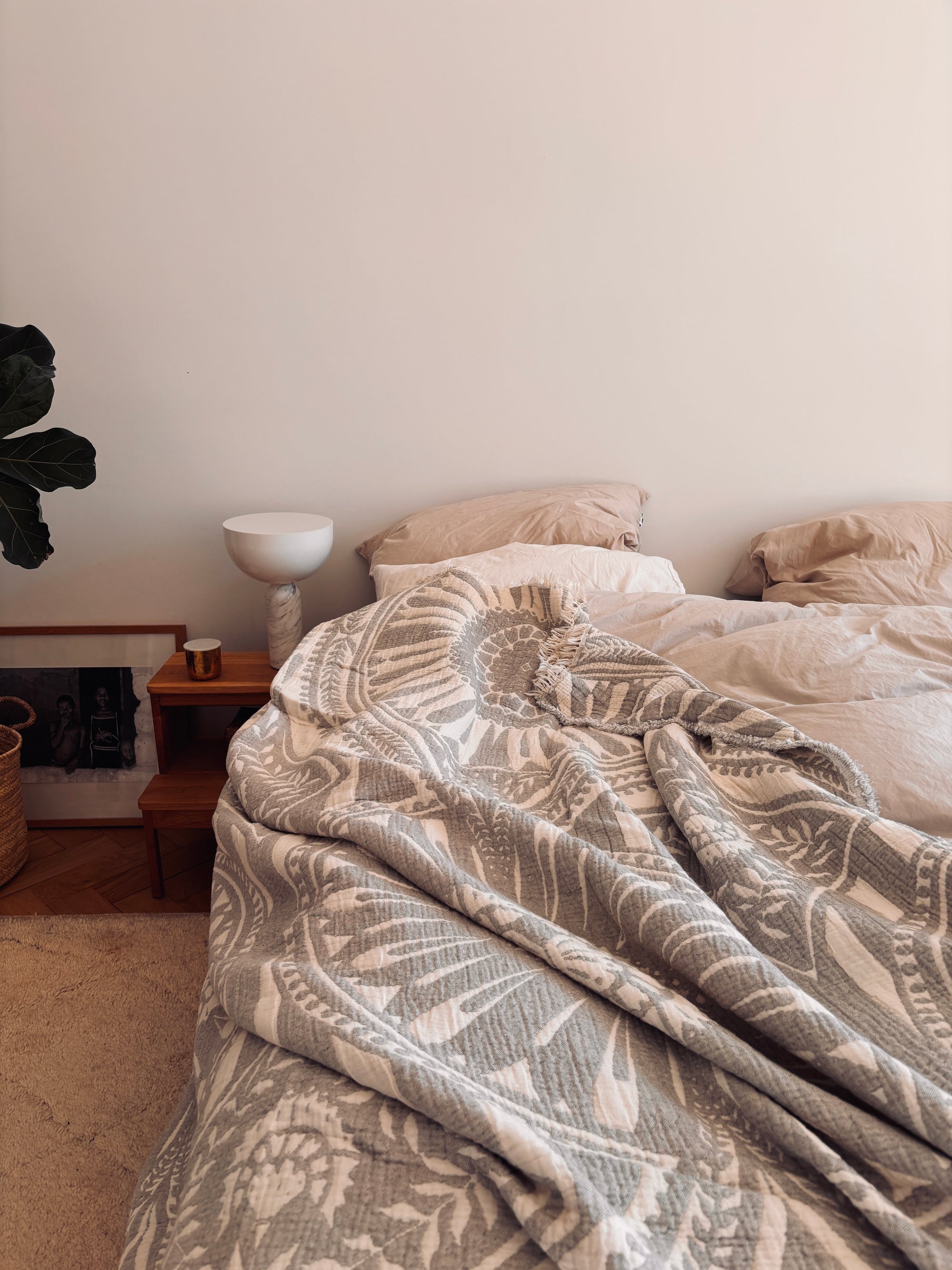 Auf einem gemütlichen, ungemachten Bett mit beiger Bettwäsche liegt die CALMA Musselin Decke in hellgrau (2,20 x 2,30 m) von LEY! und sorgt für Boho-Stil. Ein kleiner Nachttisch mit einer weißen Lampe und Dekoration steht neben gerahmter Kunst und einer Pflanze auf dem Holzboden.