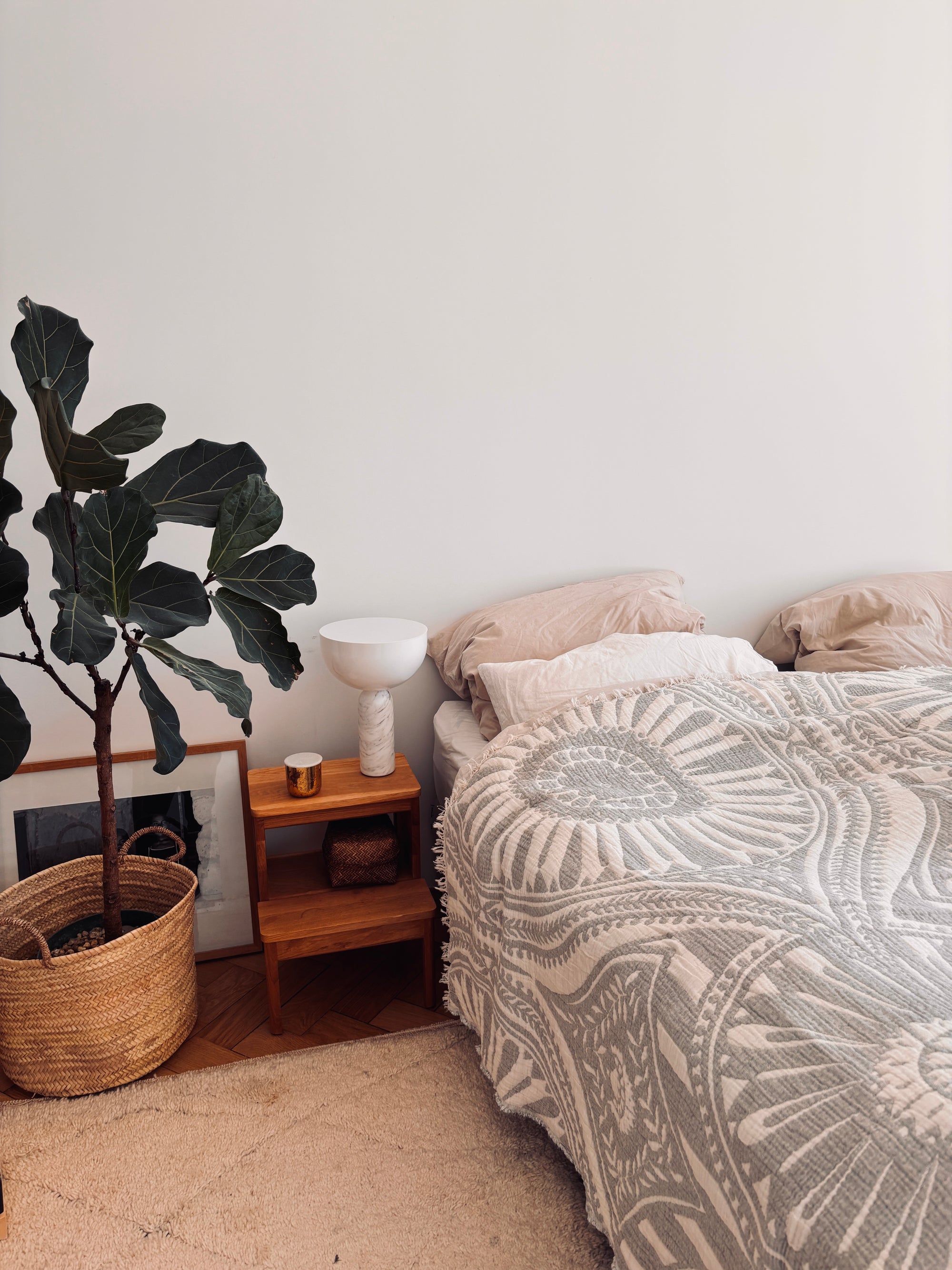 Ein gemütliches Schlafzimmer mit Boho-Design-Akzenten: die LEY! CALMA Musselin Decke in Hellgrau (2,20 x 2,30 m) auf dem Bett, beige Kissen, ein hölzerner Nachttisch mit Lampe und Kerze und eine große Topfpflanze in einem geflochtenen Korb auf dem Boden.