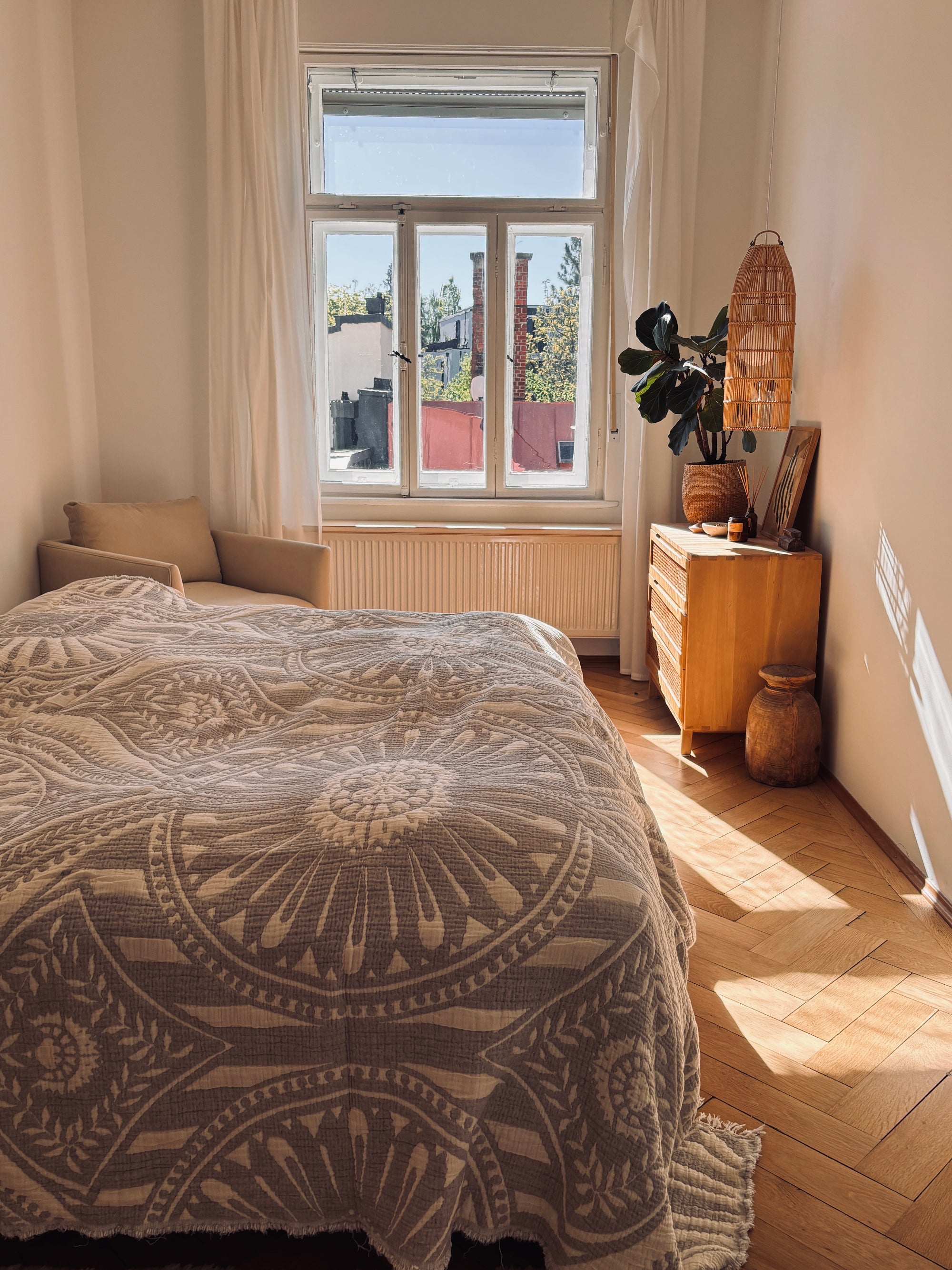 Ein sonnendurchflutetes Schlafzimmer mit Boho-Akzenten verfügt über eine LEY! CALMA Musselin Decke in Hellgrau (2,20 x 2,30 m), einem beigen Sofa, einer Holzkommode, einer Topfpflanze und dem Blick auf die Dachterrasse. Warmes Licht wirft weiche Schatten auf den atmungsaktiven Holzboden.