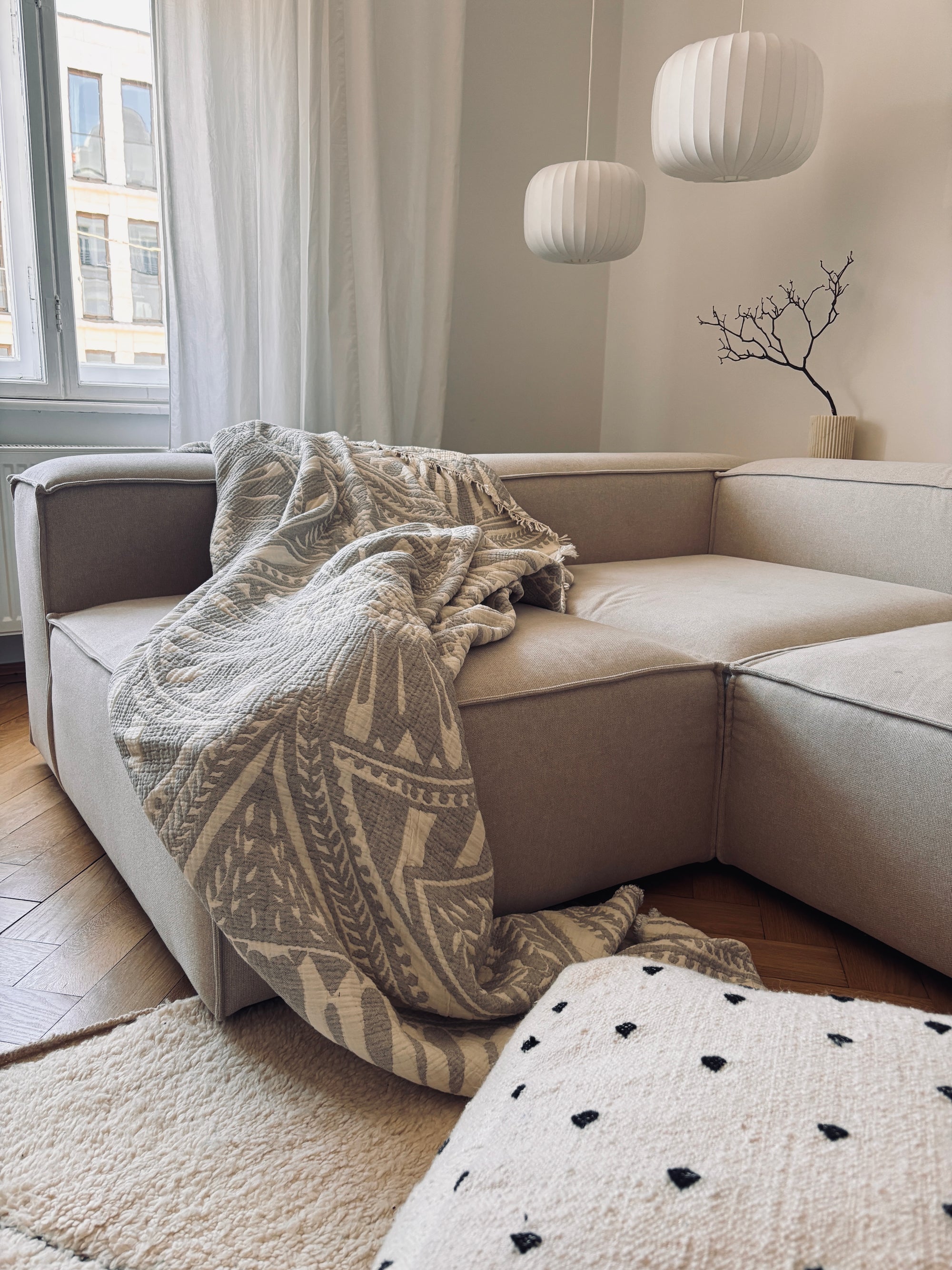 Eine gemütliche beigefarbene Couchgarnitur mit der LEY! CALMA Musselin Decke in Hellgrau (2,20 x 2,30 m), die darüber drapiert ist, sitzt an einem Fenster mit transparenten, atmungsaktiven Vorhängen. Moderne Pendelleuchten und ein Vasenast vervollständigen das entspannte Boho-Design-Ambiente.