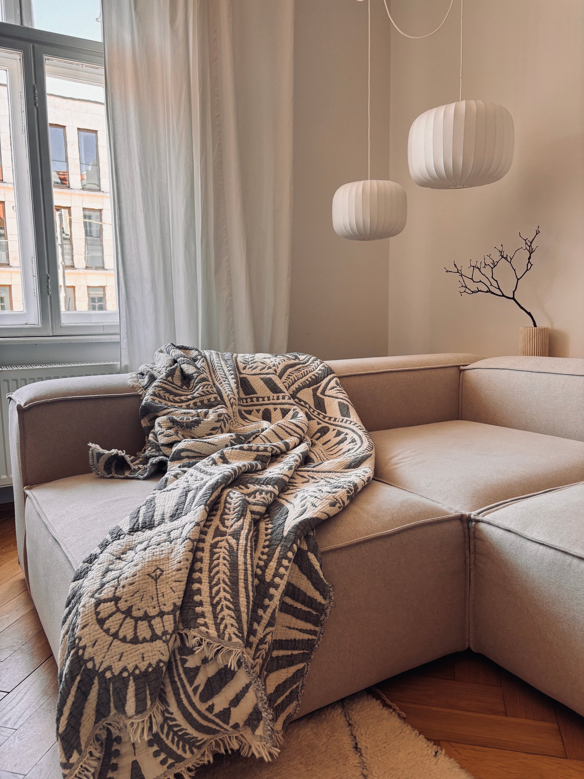 Ein gemütliches beigefarbenes Sektionssofa, über das die CALMA Musselin Decke in Dunkelgrau von LEY! drapiert ist, neben einem großen Fenster mit transparenten Vorhängen. Darüber hängen drei weiße Pendelleuchten, und eine minimalistische Vase mit Zweigen unterstreicht das Boho-Design.