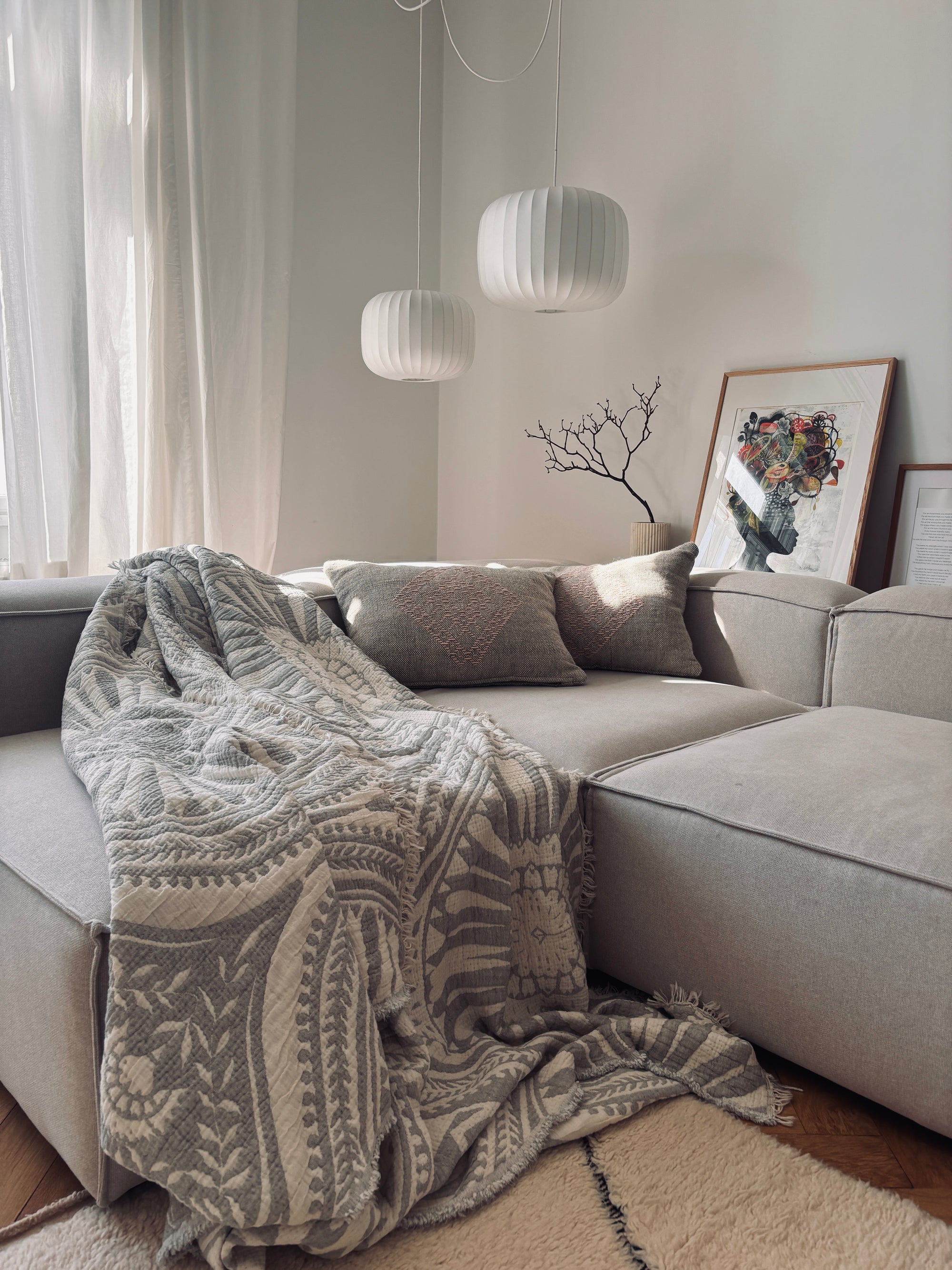 Ein gemütliches Wohnzimmer mit einem hellgrauen Sektionssofa, LEY! CALMA Musselin Decke in Hellgrau (2,20 x 2,30 m), dekorative Kissen, moderne weiße Pendelleuchten, durchsichtige Vorhänge für eine stimmungsvolle Atmosphäre und gerahmte Kunst neben einer mit Zweigen gefüllten Vase.