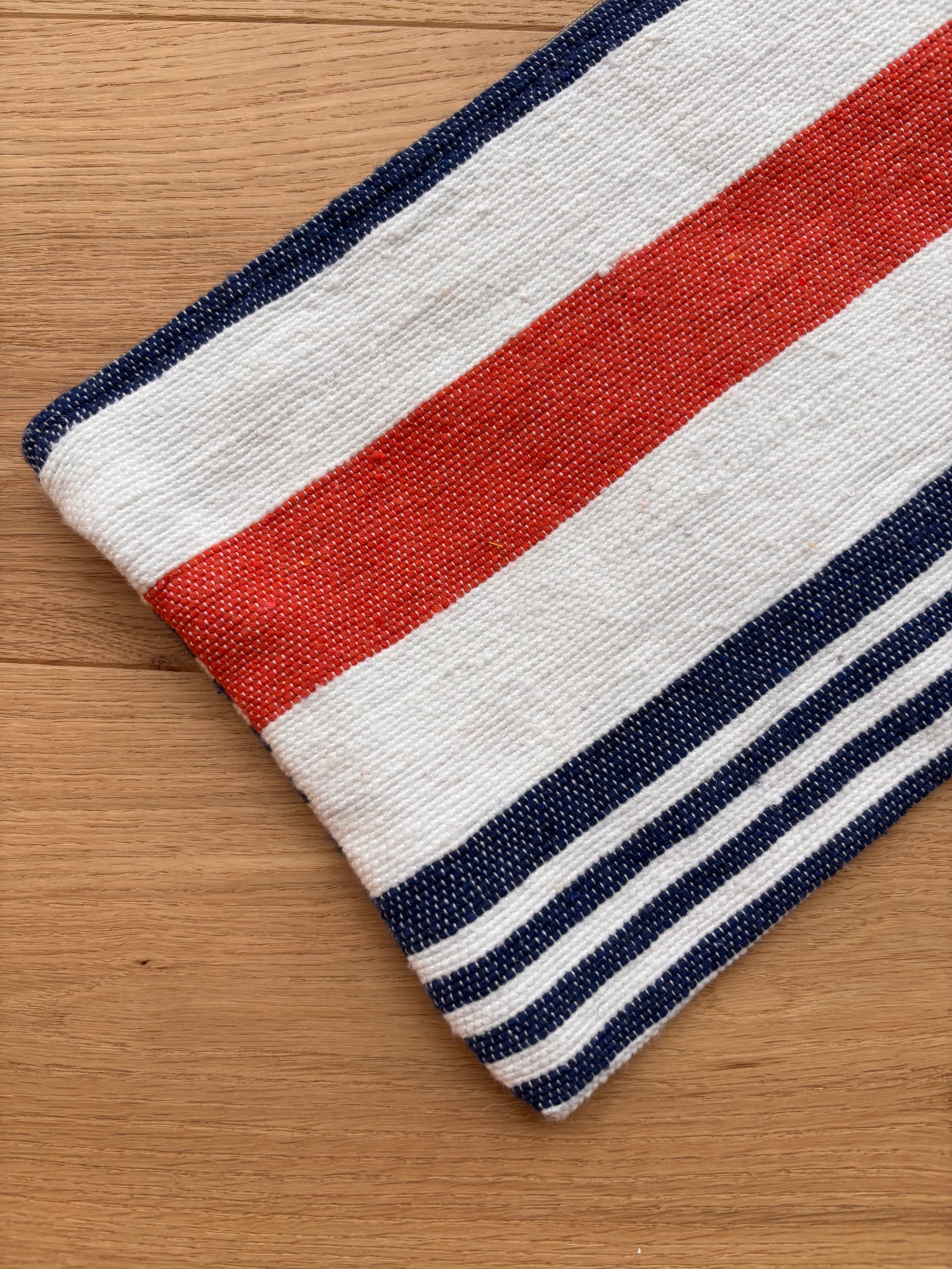 Ein gefaltetes Tuch aus der Tasche - Stripe Line "Parosa" (20x30cm) von LEY! mit kräftigen roten, weißen und blauen Streifen ruht auf Holz - ein idealer stilvoller Akzent für Ihre Baumwolltasche.