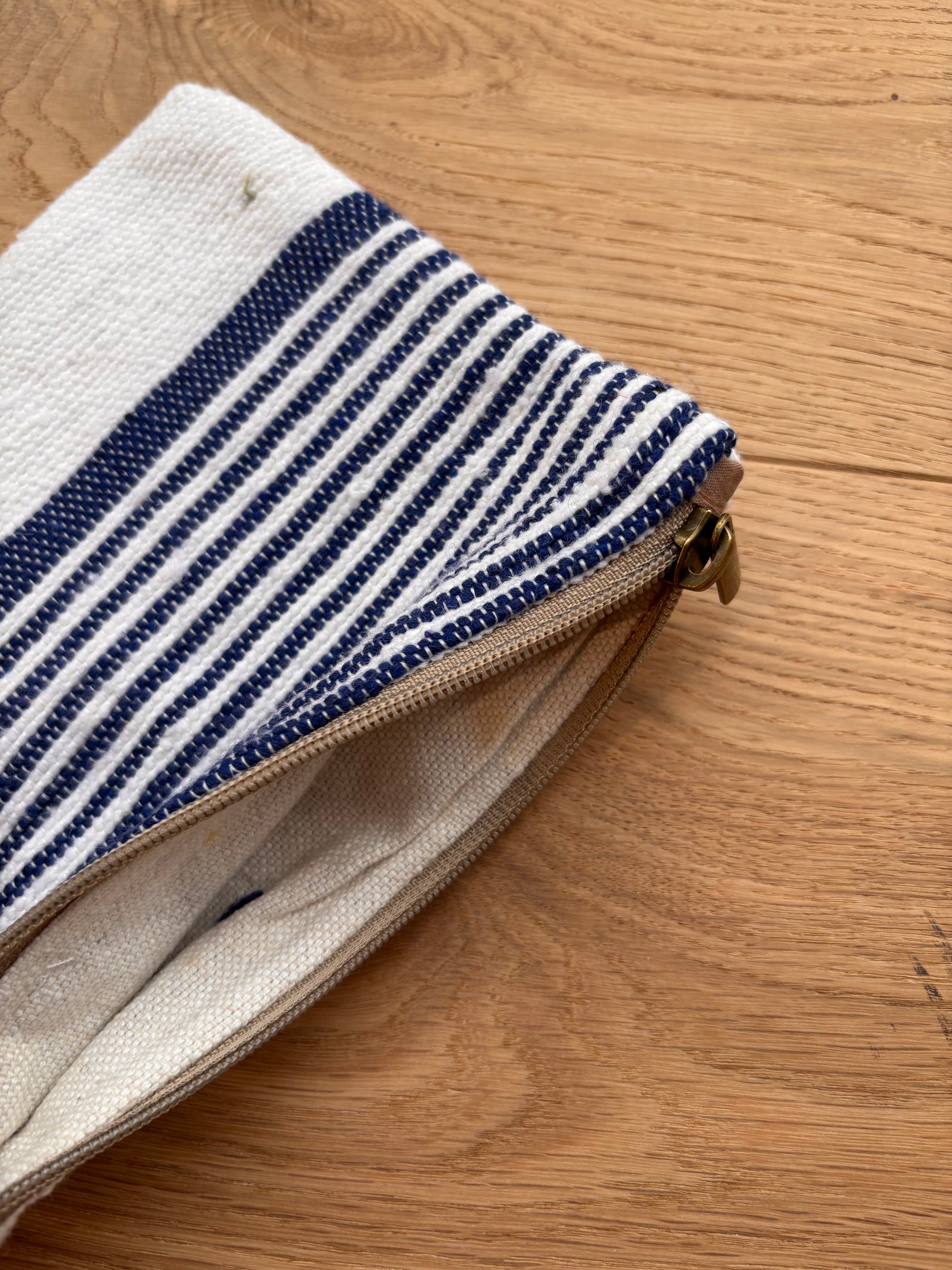 Eine Nahaufnahme der LEY! Tasche - Stripe Line "Parosa" (20x30cm) mit blauen und weißen Streifen und einem hellbraunen Reißverschluss mit Metallzug liegt teilweise geöffnet auf dem Holz und zeigt ihr handwerkliches Detail.
