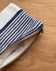 Eine Nahaufnahme der LEY! Tasche - Stripe Line "Parosa" (20x30cm) mit blauen und weißen Streifen und einem hellbraunen Reißverschluss mit Metallzug liegt teilweise geöffnet auf dem Holz und zeigt ihr handwerkliches Detail.