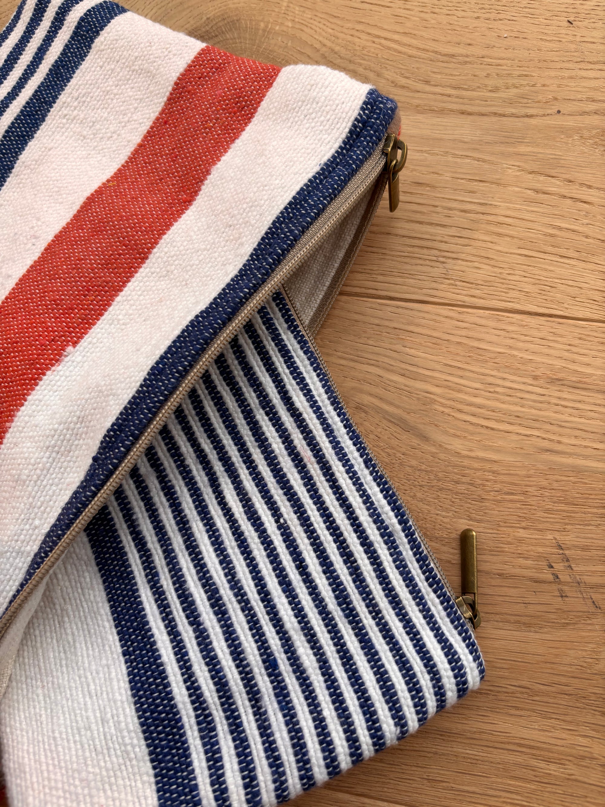 Eine Nahaufnahme der LEY! Tasche - Stripe Line "Parosa" (20x30cm), einer Baumwolltasche mit Reißverschluss, die auf der einen Seite rote und weiße Streifen und auf der anderen Seite marineblaue und weiße Streifen aufweist und auf einer hellen Holzunterlage liegt.