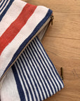 Eine Nahaufnahme der LEY! Tasche - Stripe Line "Parosa" (20x30cm), einer Baumwolltasche mit Reißverschluss, die auf der einen Seite rote und weiße Streifen und auf der anderen Seite marineblaue und weiße Streifen aufweist und auf einer hellen Holzunterlage liegt.