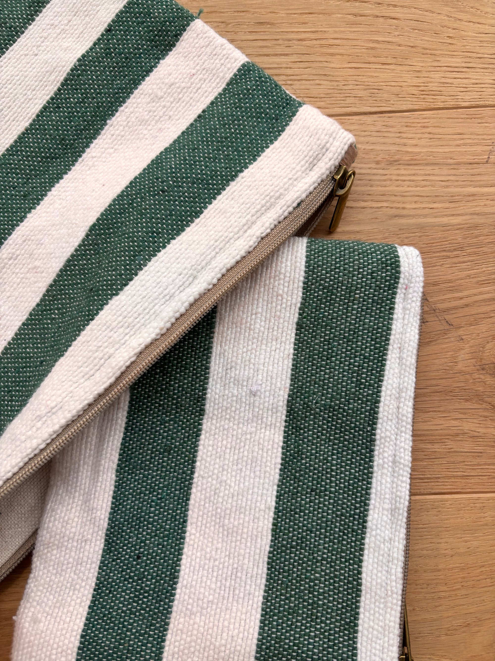 Zwei LEY! Tasche - Stripe Line "Tilosa" (20x30cm) mit grünen und cremefarbenen vertikalen Streifen und goldenen Reißverschlüssen sind überlappend auf Holz abgebildet.
