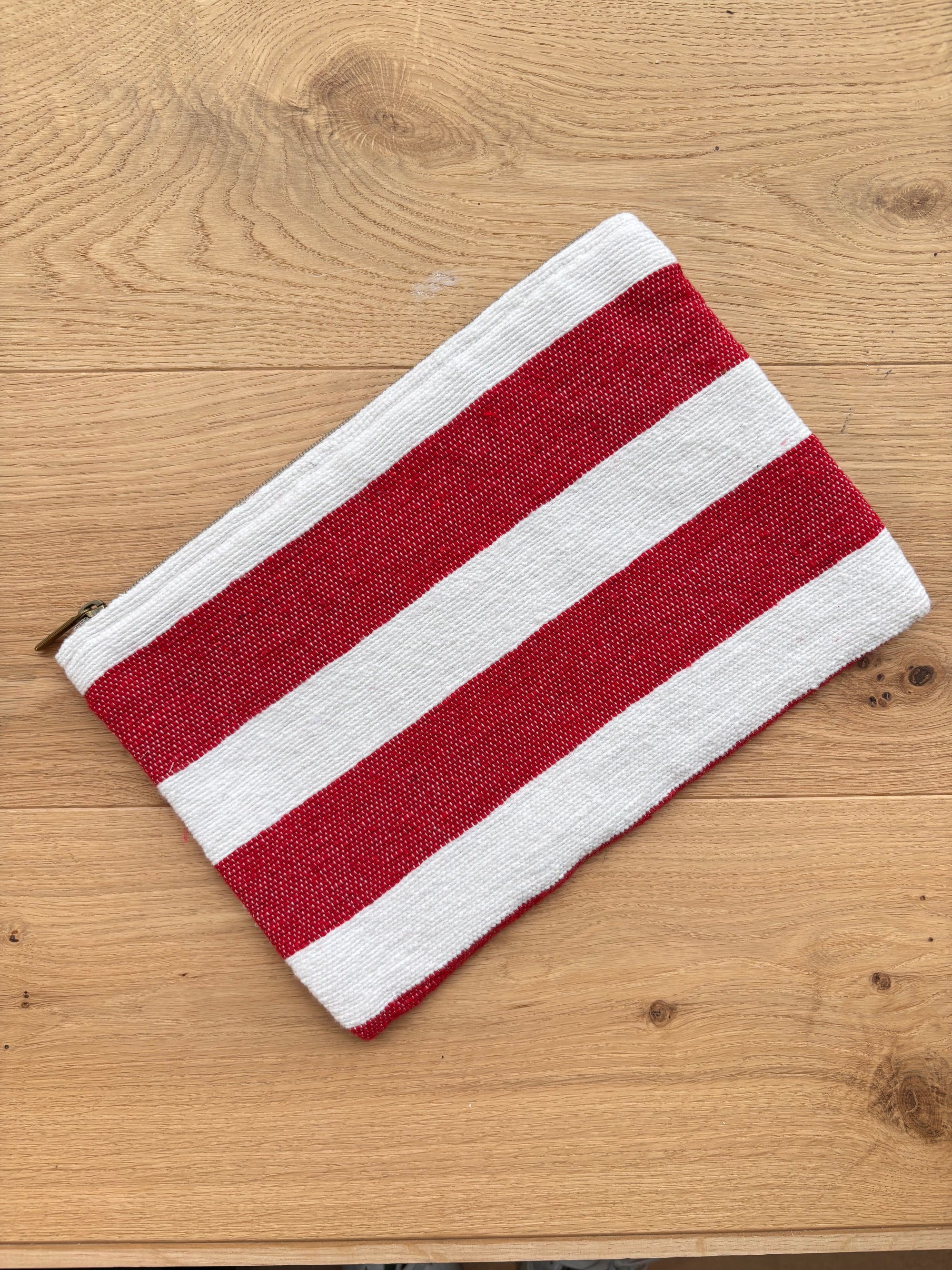 Die Tasche - Stripe Line "Symia" (20x30cm) von LEY! aus Baumwolle mit roten und weißen Streifen und Reißverschluss liegt auf hellem Holz - handgefertigte Qualität aus Marrakesch.