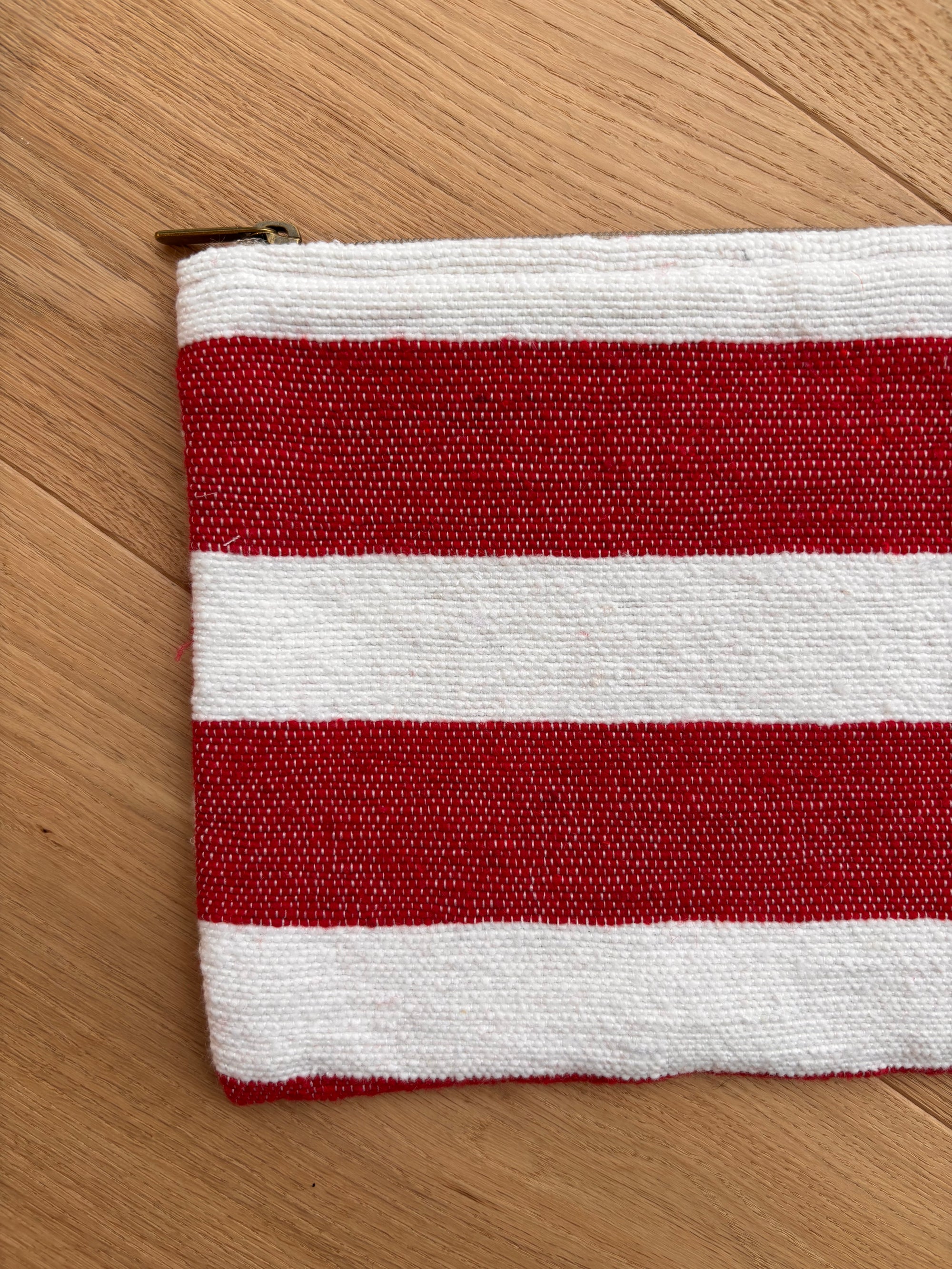 Die LEY! Tasche - Stripe Line "Symia" (20x30cm) besticht durch handgemachtes Streifendekor in Rot-Weiß und sichtbaren Reißverschluss an der linken Ecke - teils auf heller Holzfläche präsentiert.