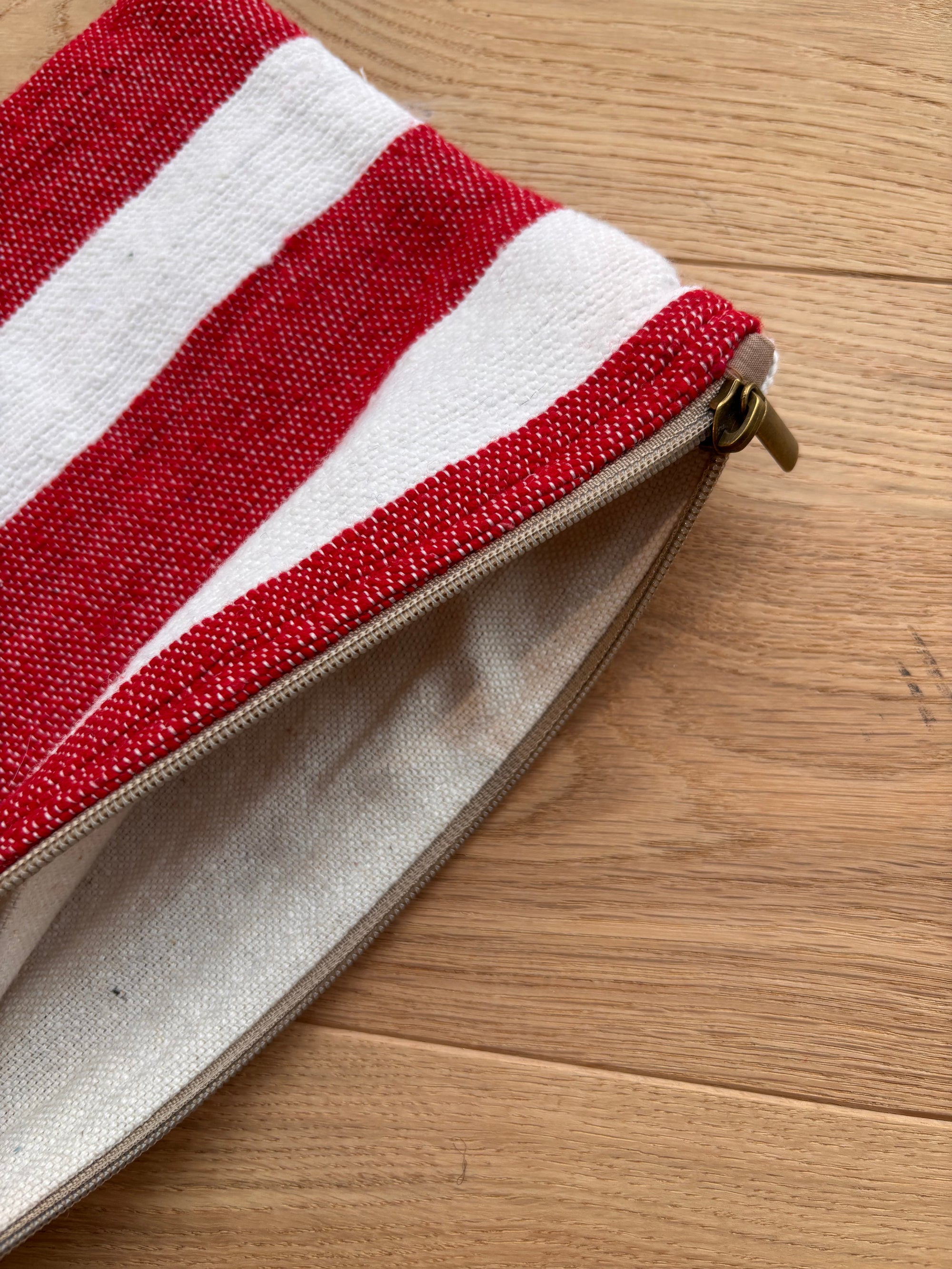 Die Tasche - Stripe Line "Symia" (20x30cm) von LEY! mit rot-weißen Streifen und beigem Reißverschluss liegt halb geöffnet auf heller Holzoberfläche.