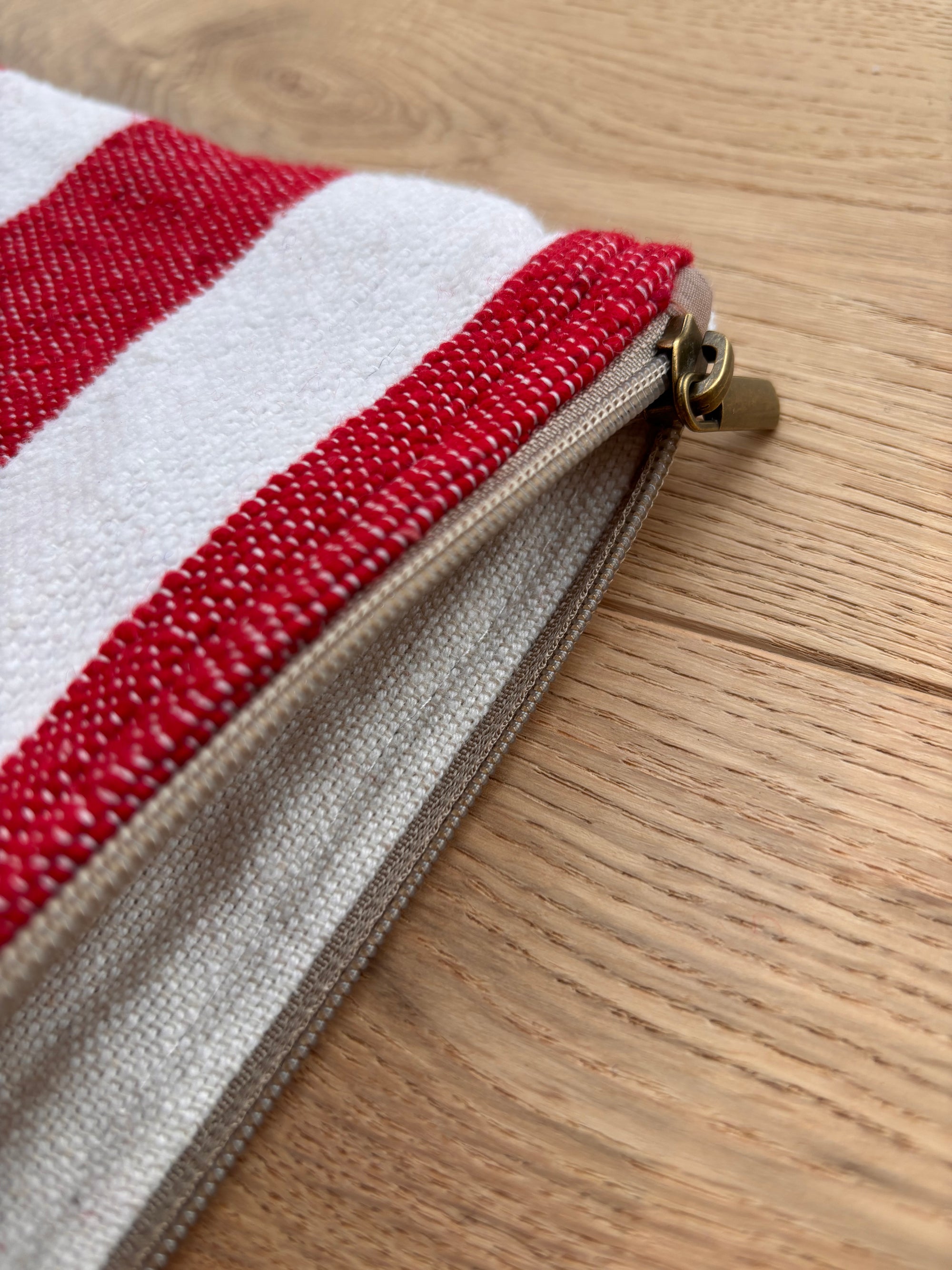Eine Nahaufnahme der LEY! Tasche - Stripe Line "Symia" (20x30cm) zeigt ihr rot-weiß gestreiftes Design und den hellbraunen Reißverschluss, der auf einer hellen Holzoberfläche liegt. Der Reißverschluss ist teilweise geöffnet und gibt den Blick auf das Stofffutter im Inneren frei.