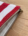Eine Nahaufnahme der LEY! Tasche - Stripe Line "Symia" (20x30cm) zeigt ihr rot-weiß gestreiftes Design und den hellbraunen Reißverschluss, der auf einer hellen Holzoberfläche liegt. Der Reißverschluss ist teilweise geöffnet und gibt den Blick auf das Stofffutter im Inneren frei.