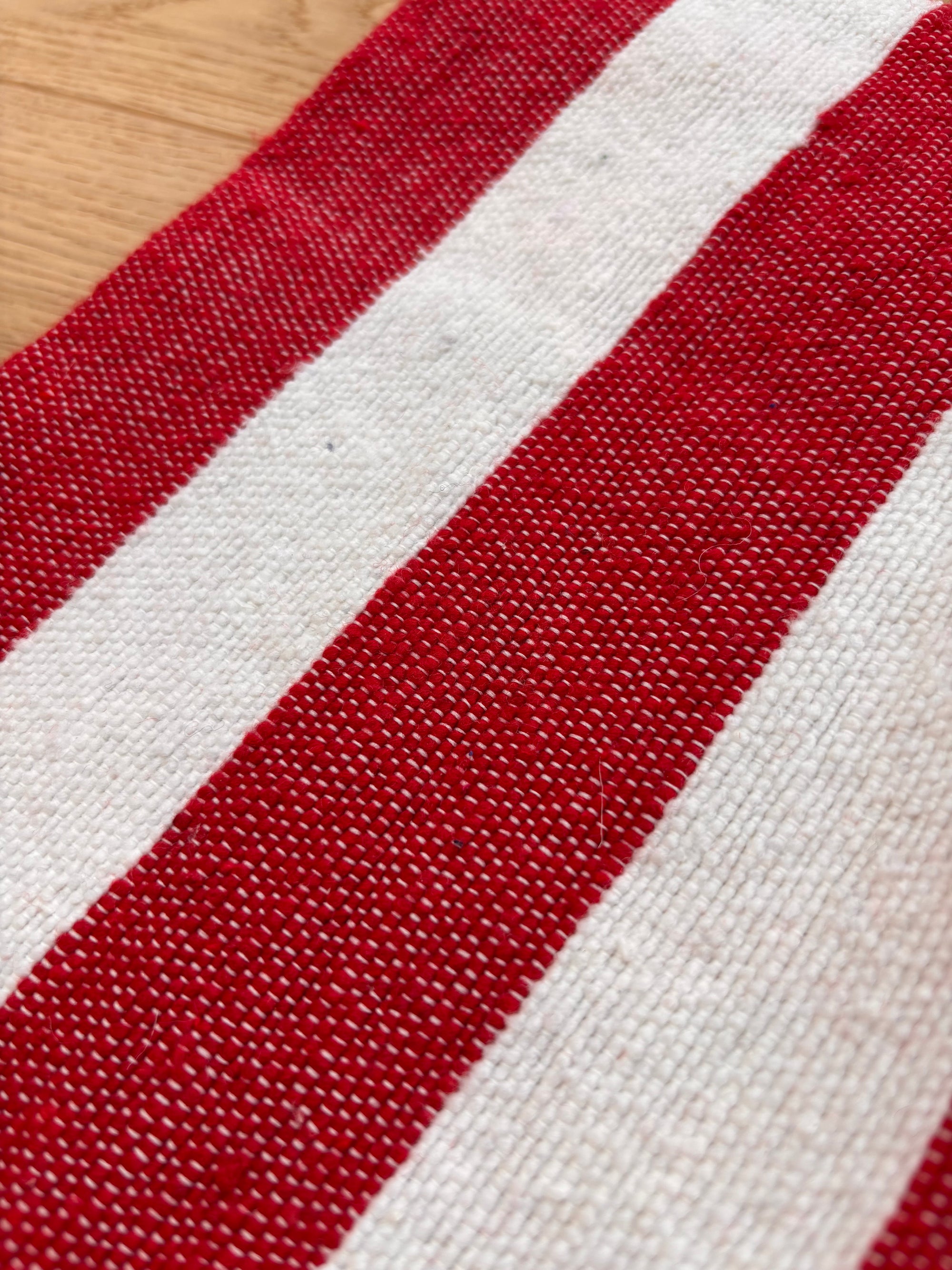 Die Nahaufnahme der LEY! Tasche - Stripe Line "Symia" (20x30cm) zeigt den gewebten Stoff mit abwechselnd roten und weißen Streifen, die die Textur und das Webmuster auf einer Holzoberfläche hervorheben.