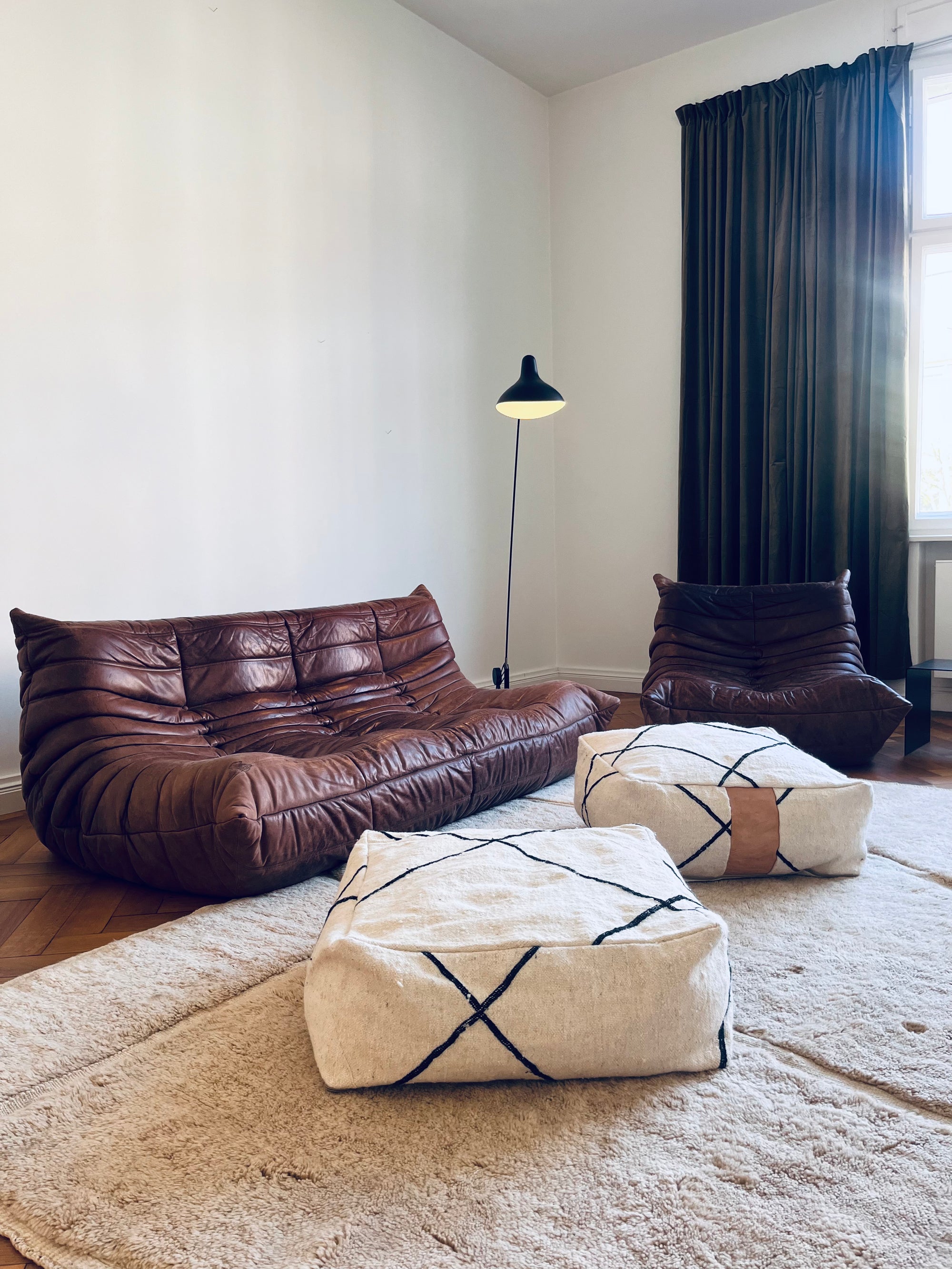 Ein minimalistisches Wohnzimmer mit einem braunen Ledersofa und -sessel, zwei weiß gemusterten Hockern, einem beigen Teppich, einer Stehlampe und dunklen Vorhängen vor einem hohen Fenster. Der Raum ist luftig und hat helle Wände.