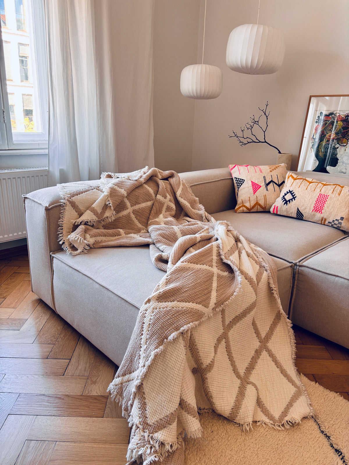 Ein beiges Sofa mit Kissen von LEY! – Colorful Line und einer Decke in einem gemütlichen, sonnendurchfluteten Wohnzimmer mit Hängeleuchten.