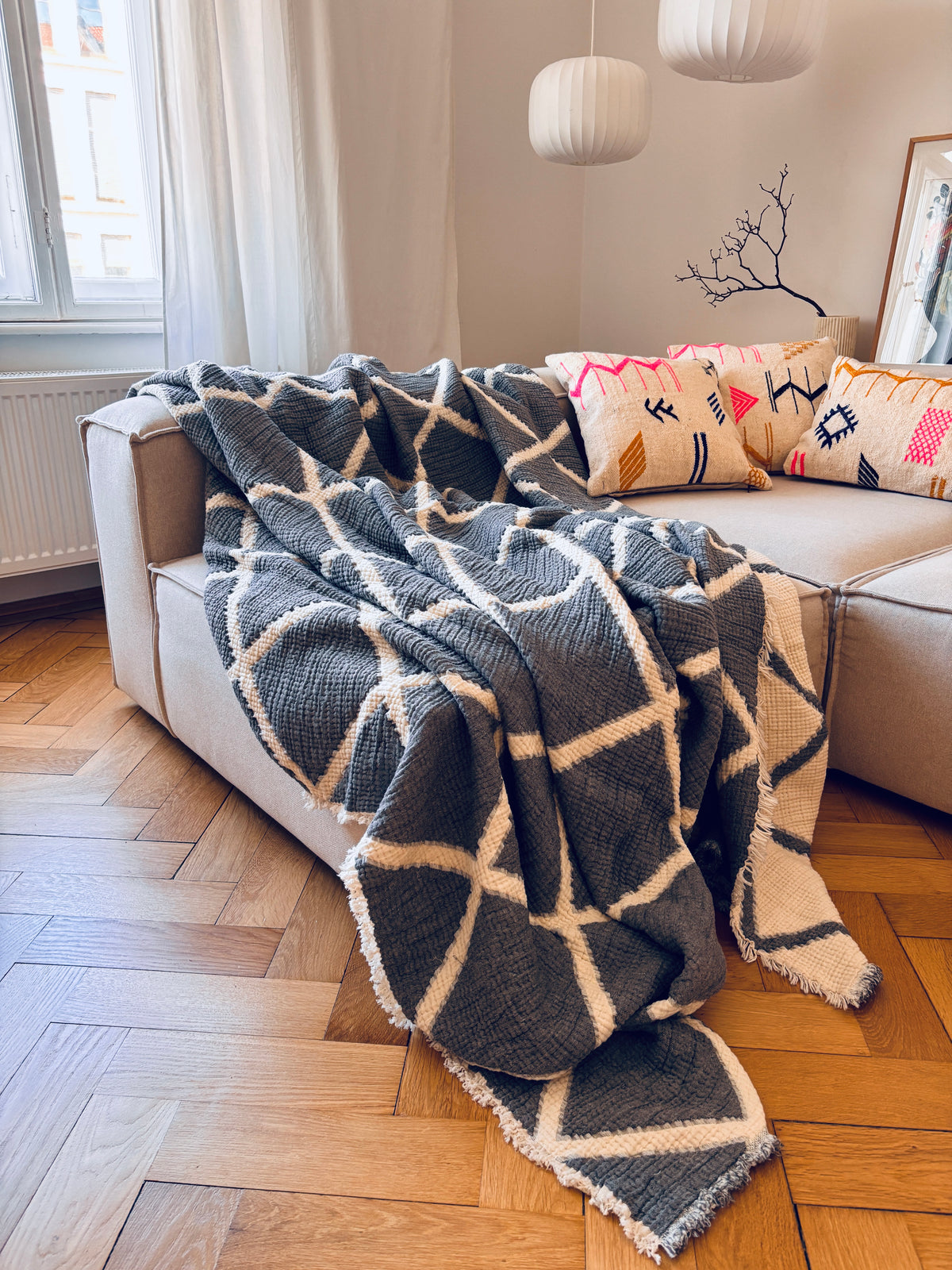 Auf einem beigen Sofa liegt eine grau-weiße Marrakesch-Decke, auf der im Sonnenlicht ein Kissen von LEY! – Colorful Line – in der Größe 40 x 60 zu sehen ist.