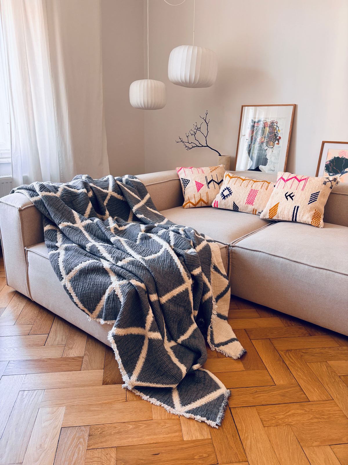 Ein beiges Sofa mit LEY!-Kissen – Colorful Line 50x50 – und einer grauen Decke in einem gemütlichen, sonnendurchfluteten Wohnzimmer.
