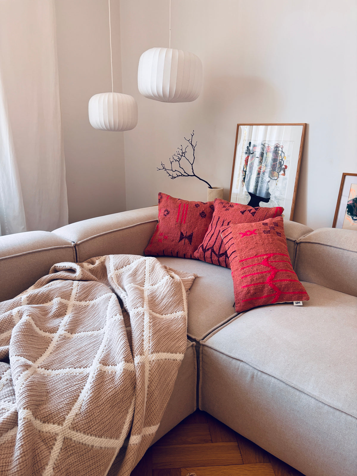 Beiges Sofa mit „LEY! Kissen Colorful Line Terracotta“ (50 x 50), einer beigen Decke und weißen Pendelleuchten darüber.