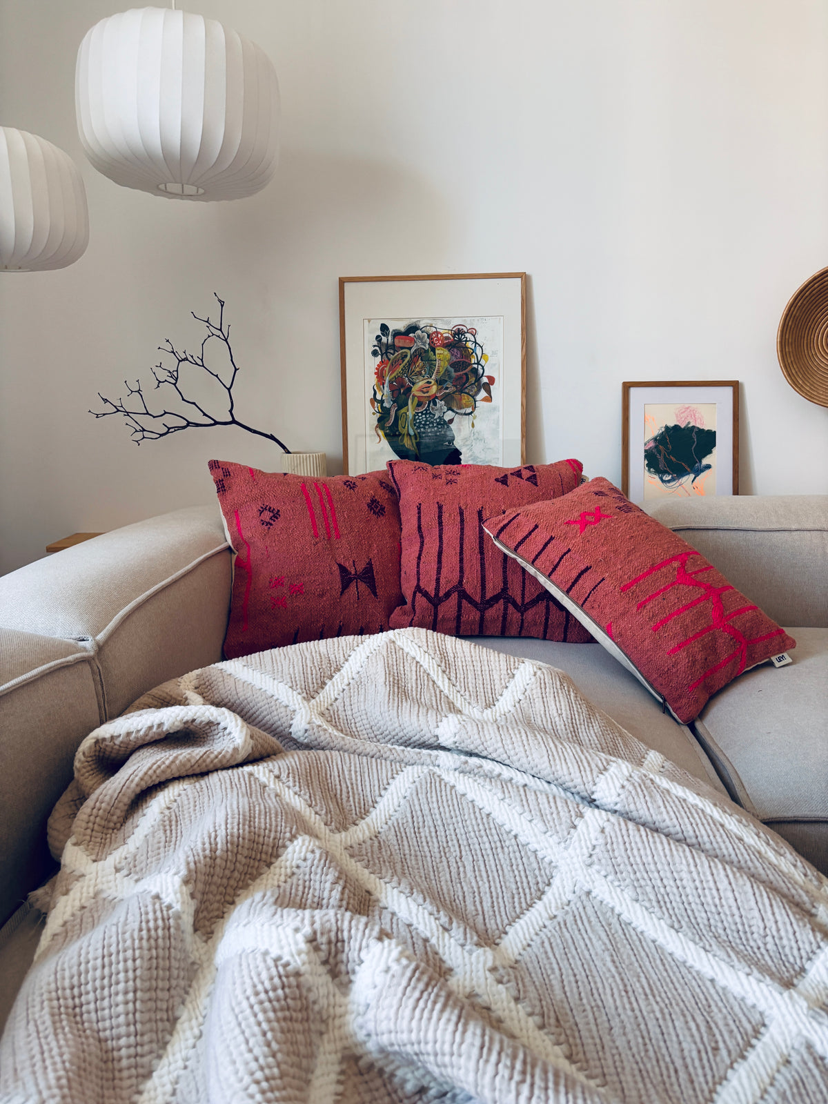 Ein gemütliches beiges Sofa mit LEY! Kissen „Colorful Line Terracotta Edition“ (50 x 50), einer cremefarbenen Strickdecke, Drucken und weißen Lampen.