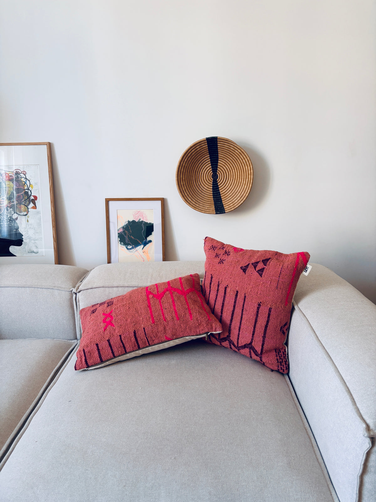 Zwei LEY!-Kissen – Colorful Line – Terracotta Edition, 50 x 50, auf einem hellen Sofa, Kunstwerk und Korb im Hintergrund.