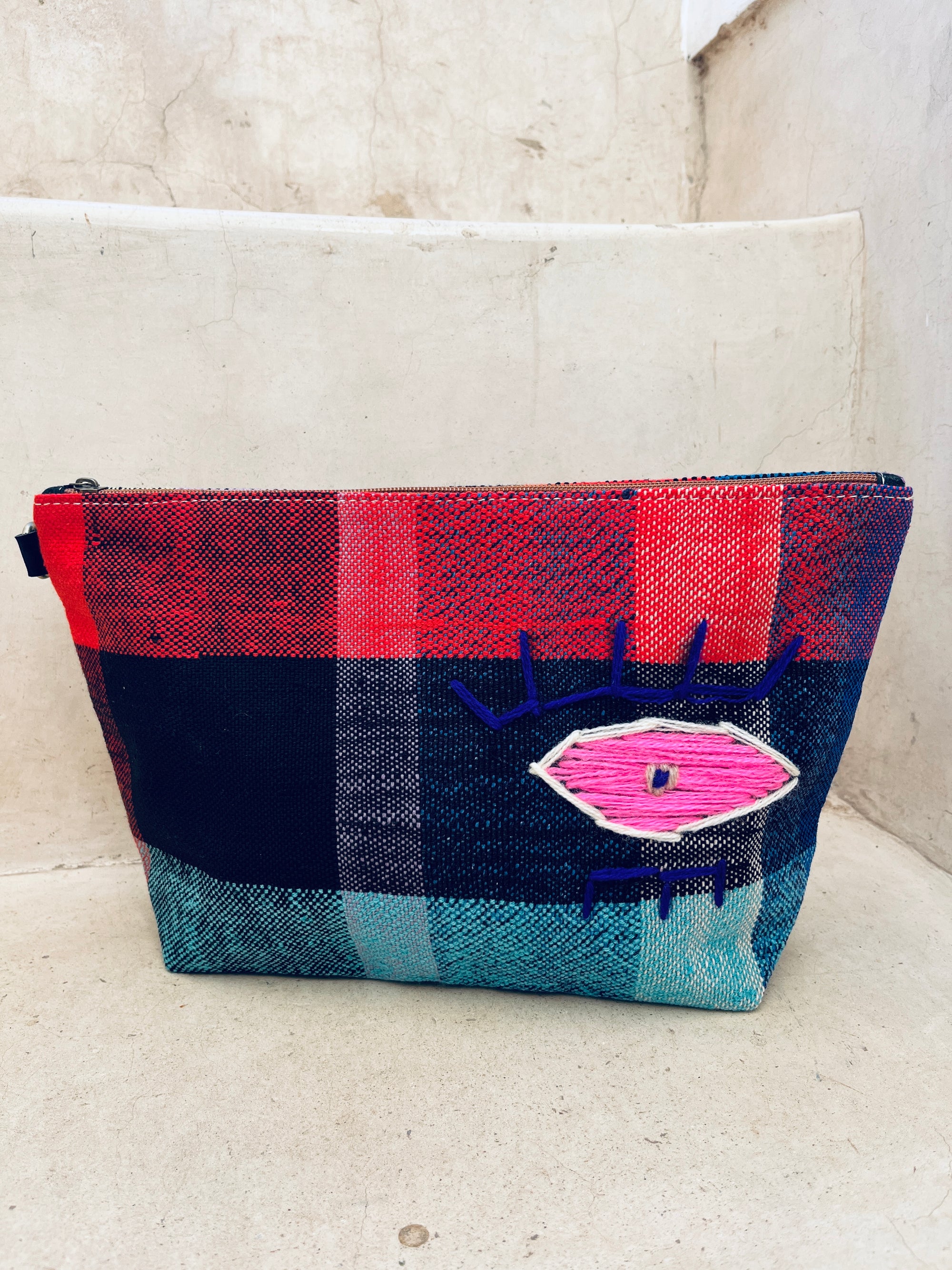 Die LEY! Tasche - Kosmetiktasche zeigt ein farbenfrohes Karomuster in Rot, Blau und Petrol mit einem gestickten rosa Auge, das auf Betonstufen vor einer beigen Wand steht.