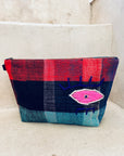 Die LEY! Tasche - Kosmetiktasche zeigt ein farbenfrohes Karomuster in Rot, Blau und Petrol mit einem gestickten rosa Auge, das auf Betonstufen vor einer beigen Wand steht.