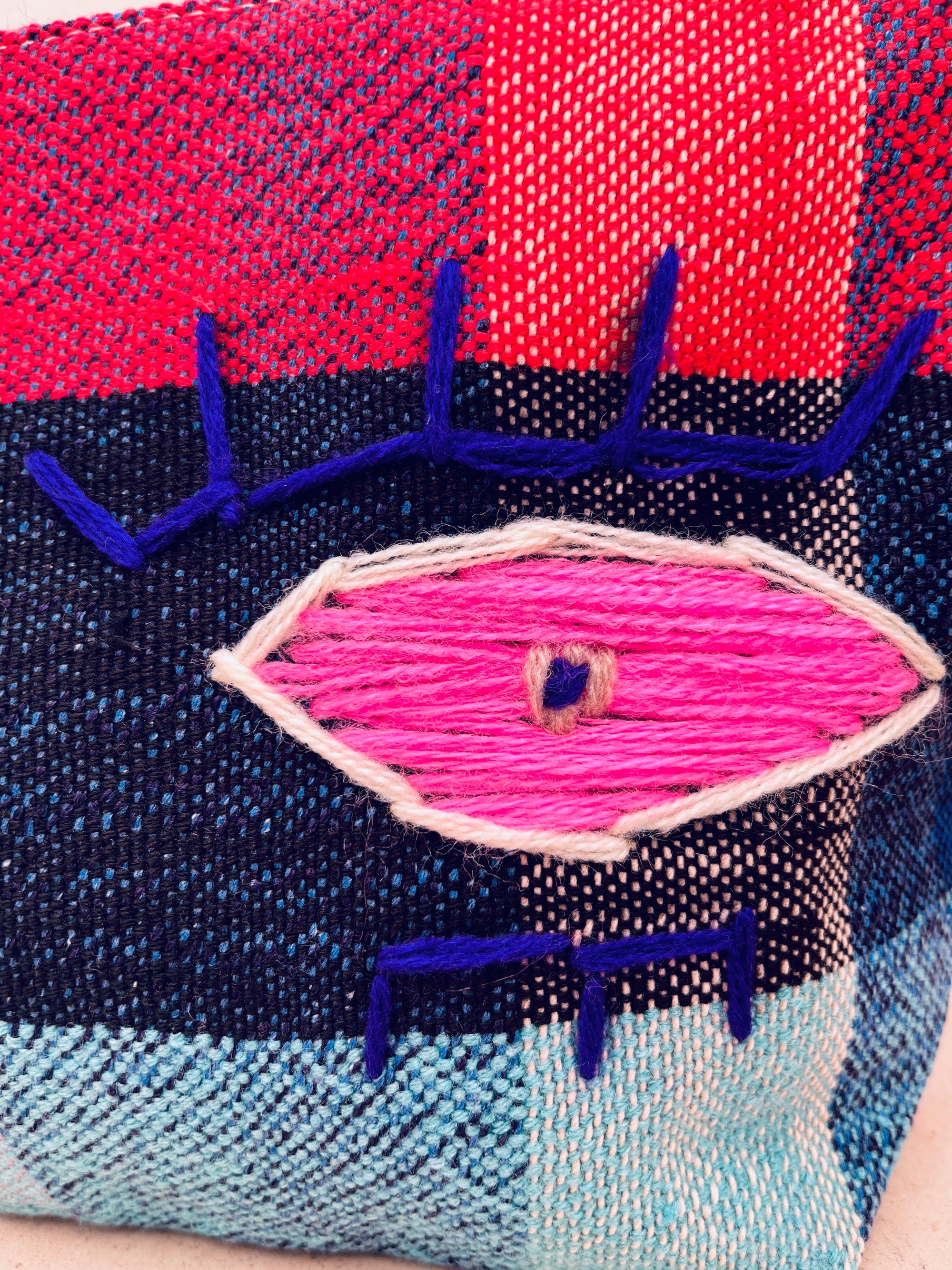 Eine Nahaufnahme der LEY! Tasche - Kosmetiktasche zeigt einen farbenfrohen, handgewebten marokkanischen Stoff mit einem gestickten Auge in Rosa, Weiß und Blau auf einem rot, marineblau und hellblau karierten Hintergrund.