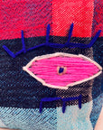 Eine Nahaufnahme der LEY! Tasche - Kosmetiktasche zeigt einen farbenfrohen, handgewebten marokkanischen Stoff mit einem gestickten Auge in Rosa, Weiß und Blau auf einem rot, marineblau und hellblau karierten Hintergrund.