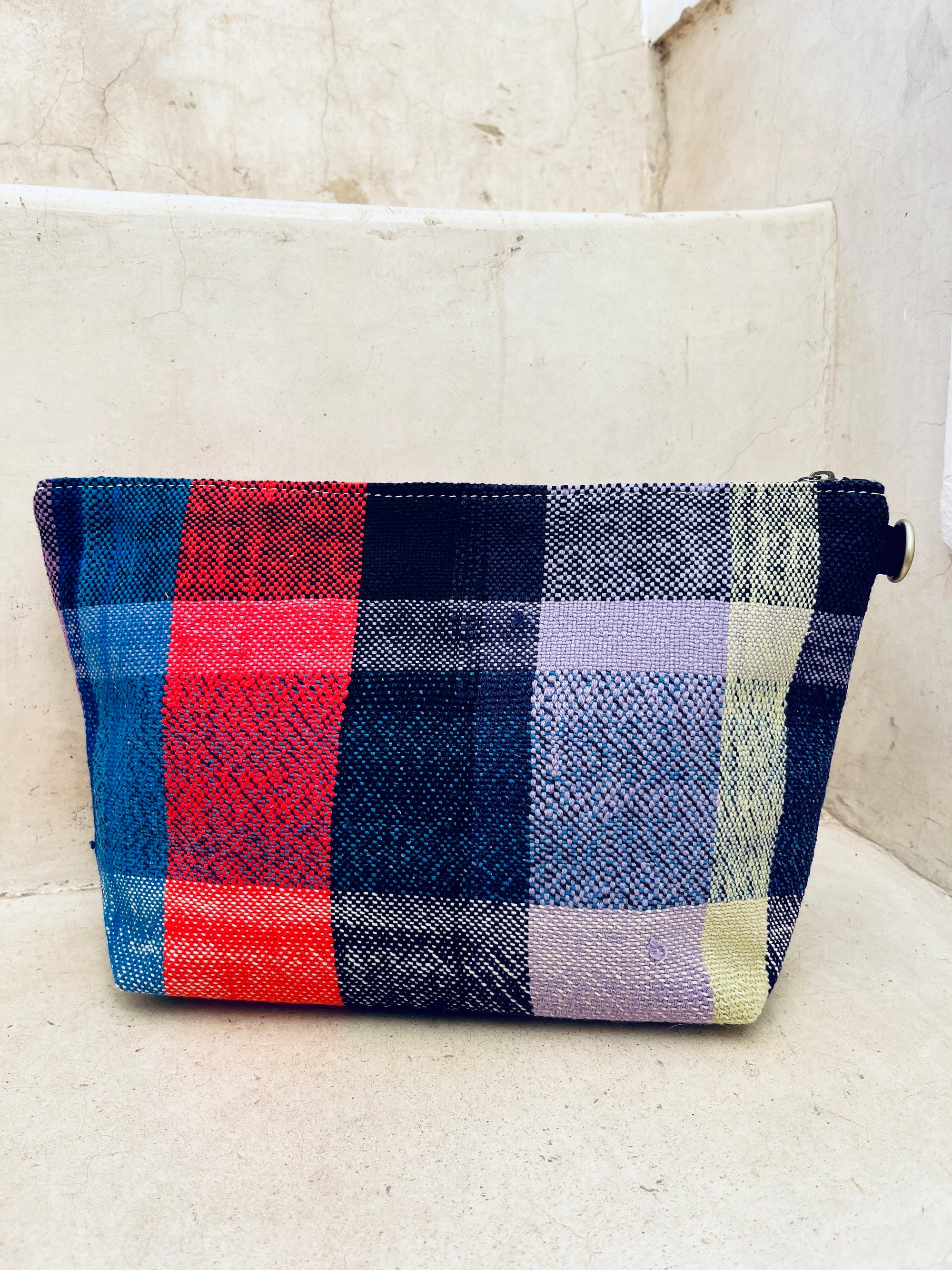 Die Tasche - Kosmetiktasche von LEY! ist handgewebt, rechteckig und hat einen Reißverschluss sowie ein buntes Karomuster in Rot, Blau, Schwarz, Gelb und Lila. Handgefertigt in Marokko und auf hellen Steinstufen platziert.
