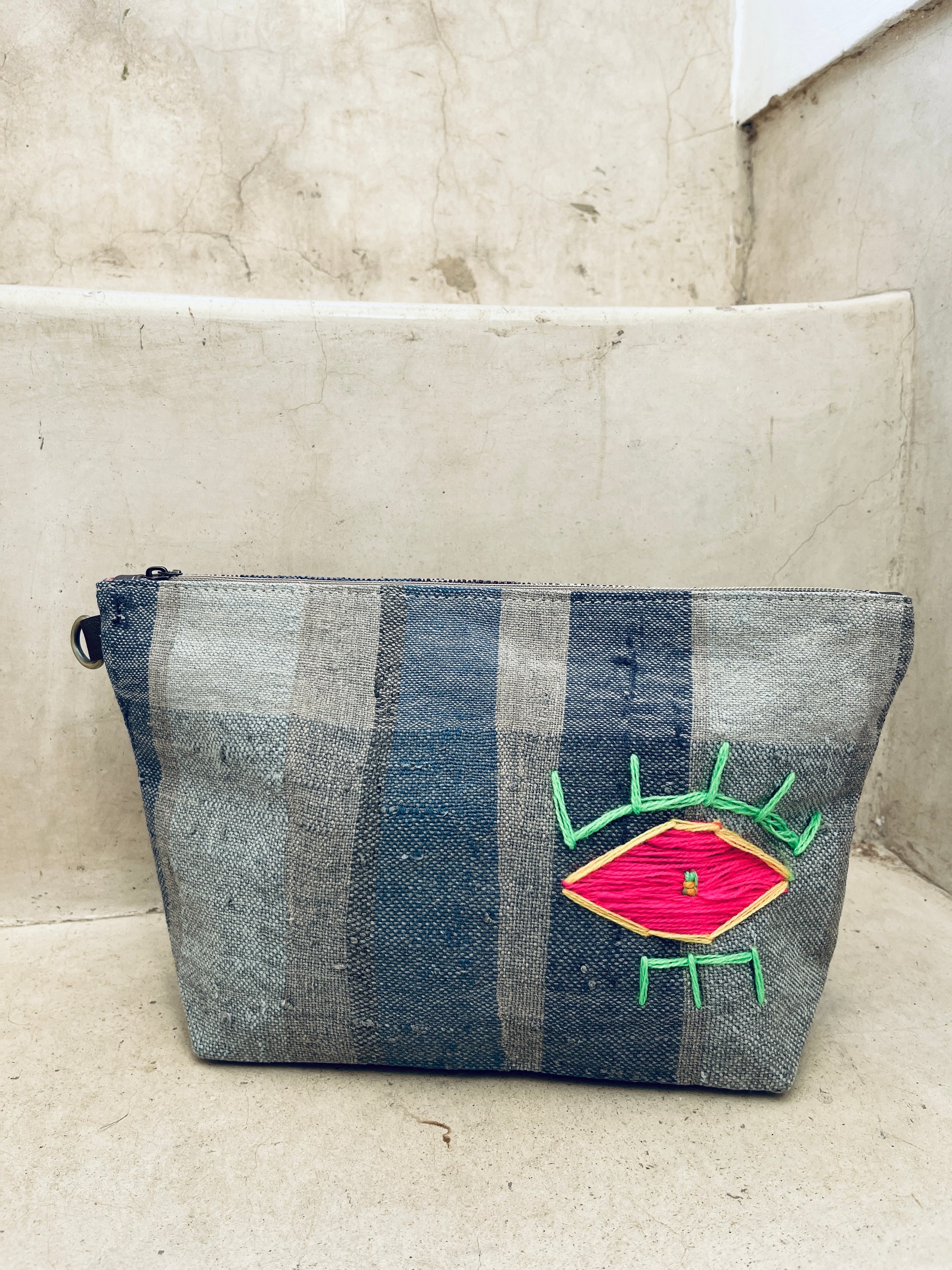 Die Tasche - Kosmetiktasche von LEY! ist grau, handgefertigt, hat vertikale blaue Streifen und ein auffälliges pink-neongrünes Augenmuster - hier auf Steinstufen präsentiert.