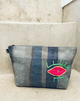 Die Tasche - Kosmetiktasche von LEY! ist grau, handgefertigt, hat vertikale blaue Streifen und ein auffälliges pink-neongrünes Augenmuster - hier auf Steinstufen präsentiert.