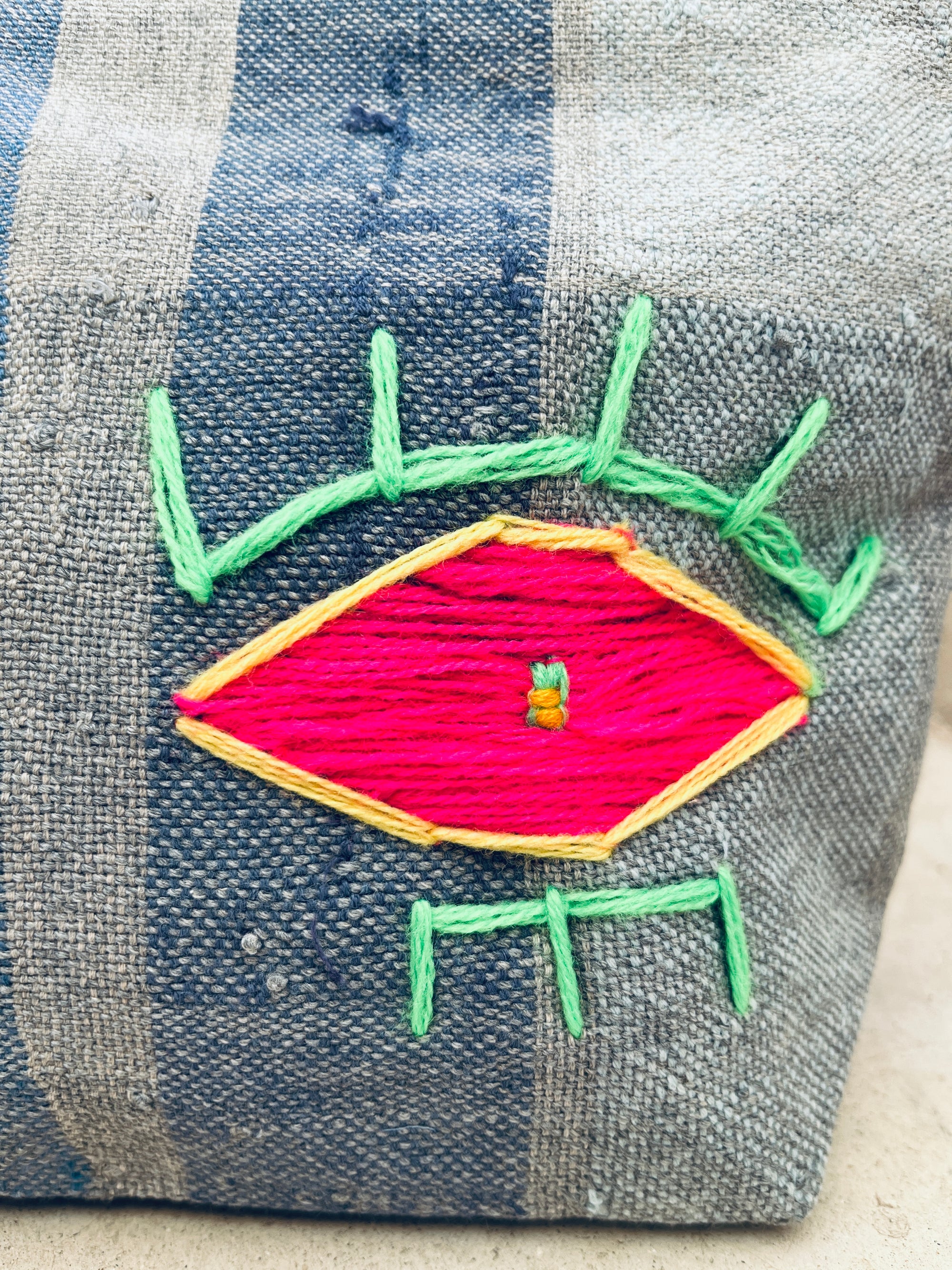 Eine Nahaufnahme des blau und grau gestreiften Stoffes der LEY! Tasche - Kosmetiktasche, mit einem bunten gestickten Auge, das gelb umrandet, mit leuchtend rosa Faden gefüllt und mit grün gestickten Wimpern akzentuiert ist.