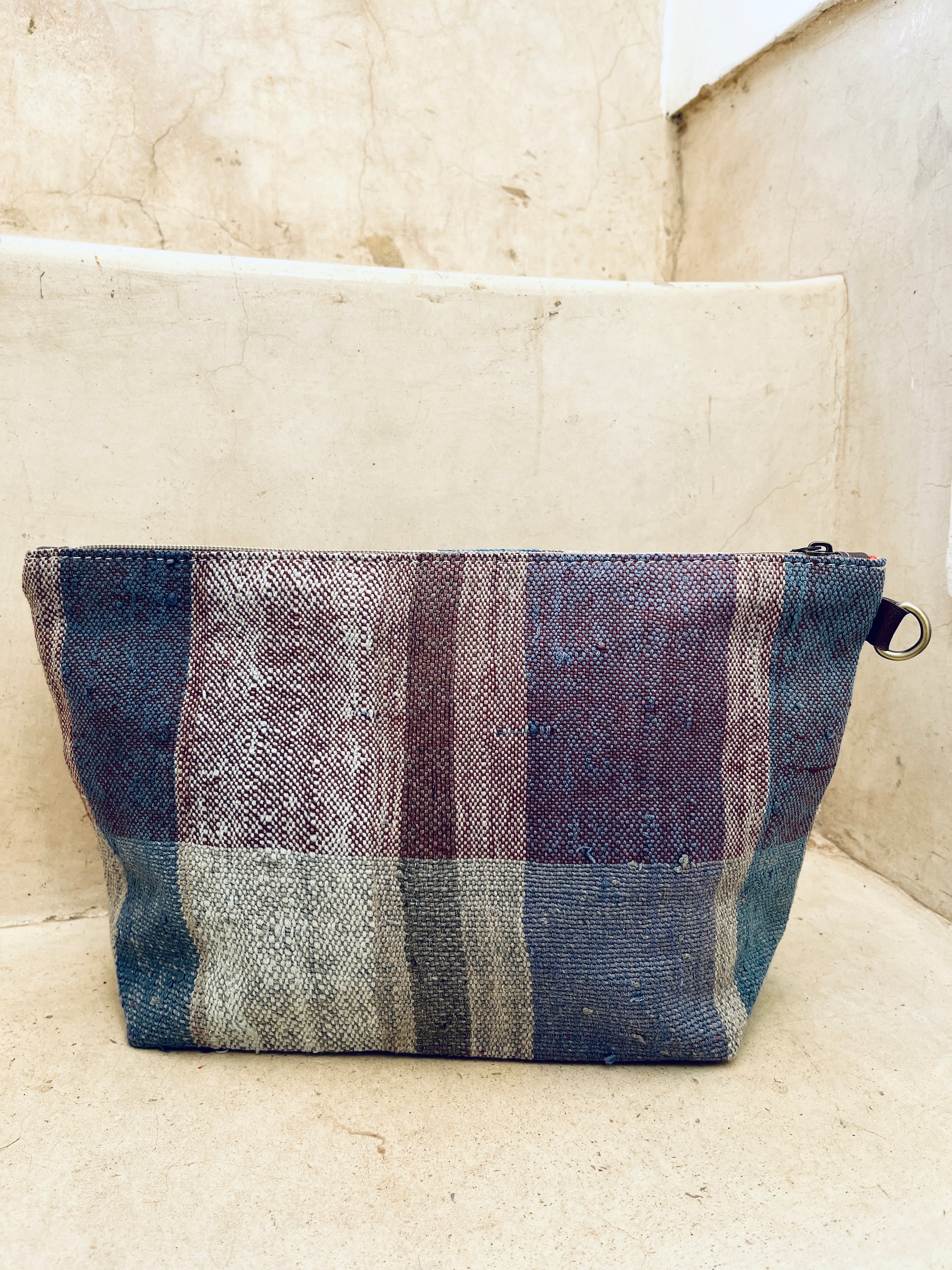 Die handgewebte Kosmetiktasche "Tasche - Kosmetiktasche" von LEY! mit Reißverschluss begeistert durch vertikale Streifen in erdigen Blau-, Lila- und Beigetönen und liegt auf einer hellen Stein- oder Betonstufe.