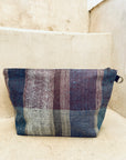 Die handgewebte Kosmetiktasche "Tasche - Kosmetiktasche" von LEY! mit Reißverschluss begeistert durch vertikale Streifen in erdigen Blau-, Lila- und Beigetönen und liegt auf einer hellen Stein- oder Betonstufe.