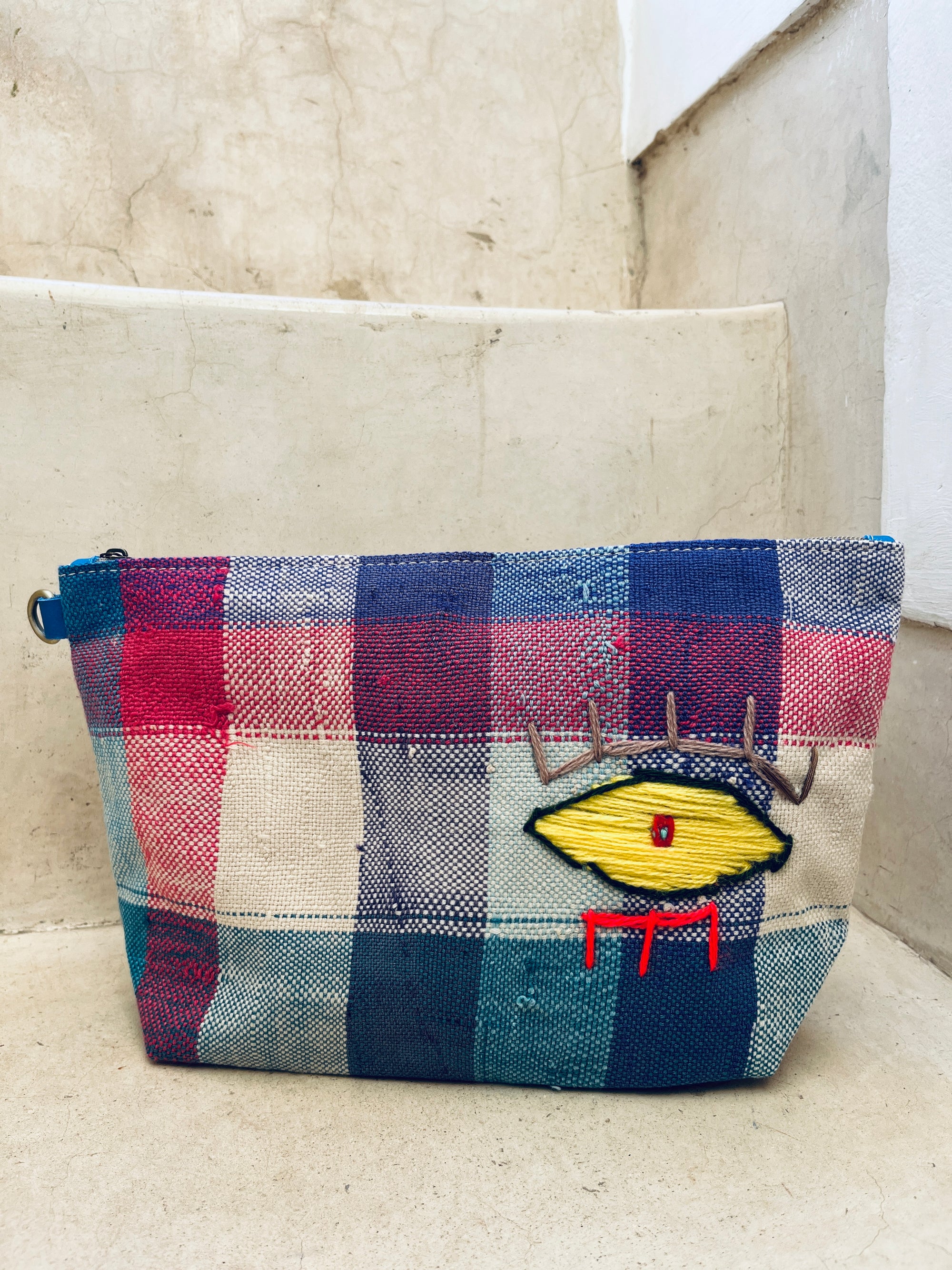 Die LEY! Kosmetiktasche besticht durch rot-weiß-blaues Karomuster, ein gesticktes gelbes Auge mit grauen Wimpern und roten Linien. Die handgefertigte kleine Tasche aus Marokko liegt auf Steinstufen.