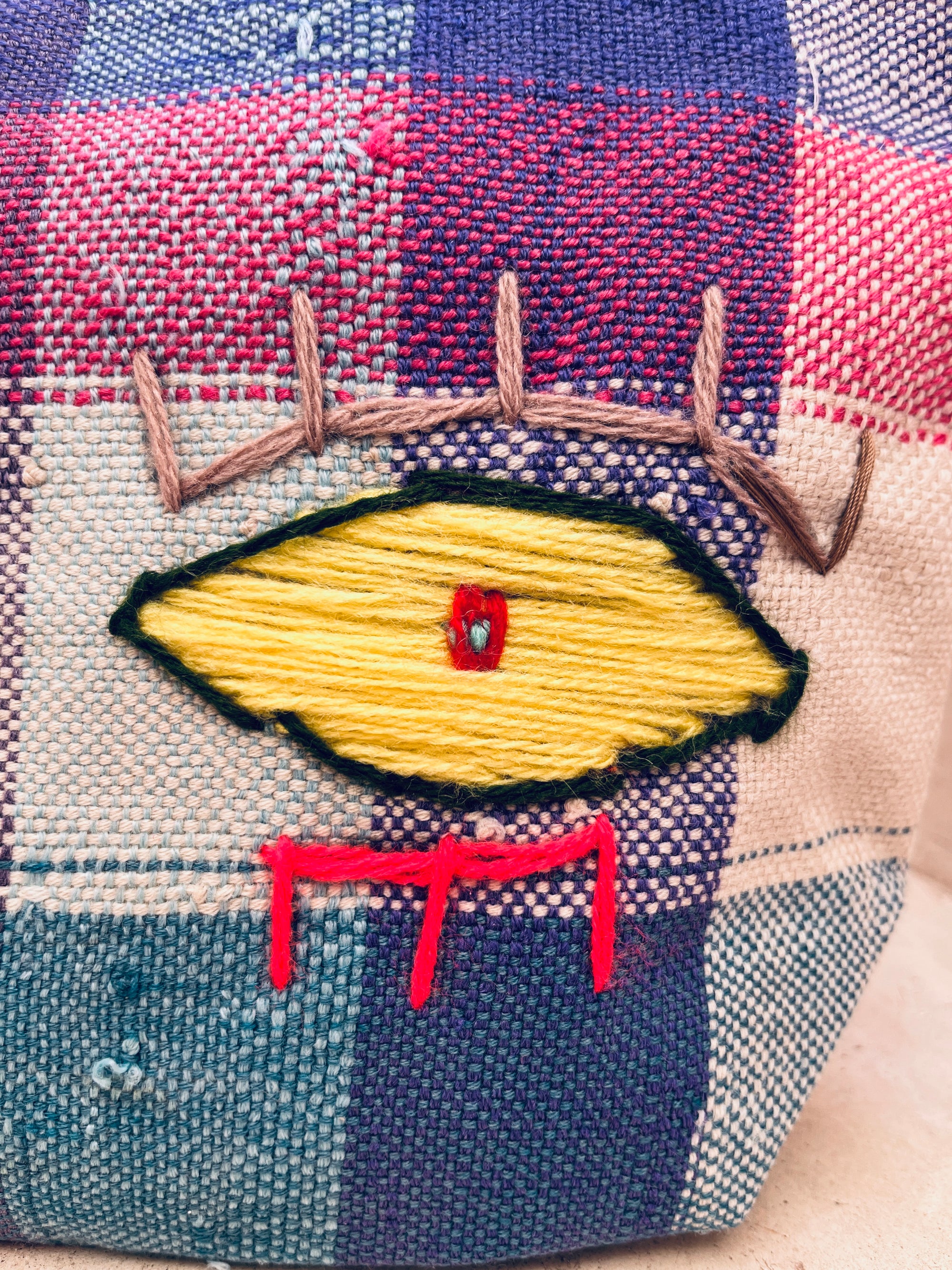 Nahaufnahme der LEY! Tasche - Kosmetiktasche, handgewebt mit einem roten, blauen und cremefarbenen Karomuster, mit einem gestickten gelben Auge mit braunen Wimpern und roten Linien darunter.