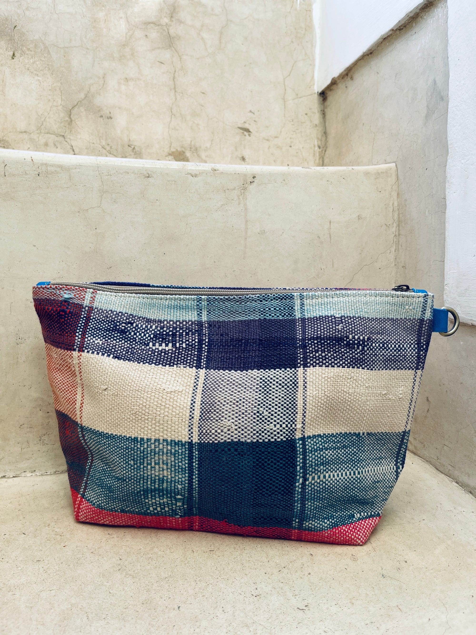 Die handgewebte Tasche - Kosmetiktasche von LEY! mit rechteckiger Form, Plaidmuster in Blau, Rot, Creme und Beige sowie Reißverschluss liegt auf einer Betontreppe vor strukturierter Wand. Hergestellt in Marokko.